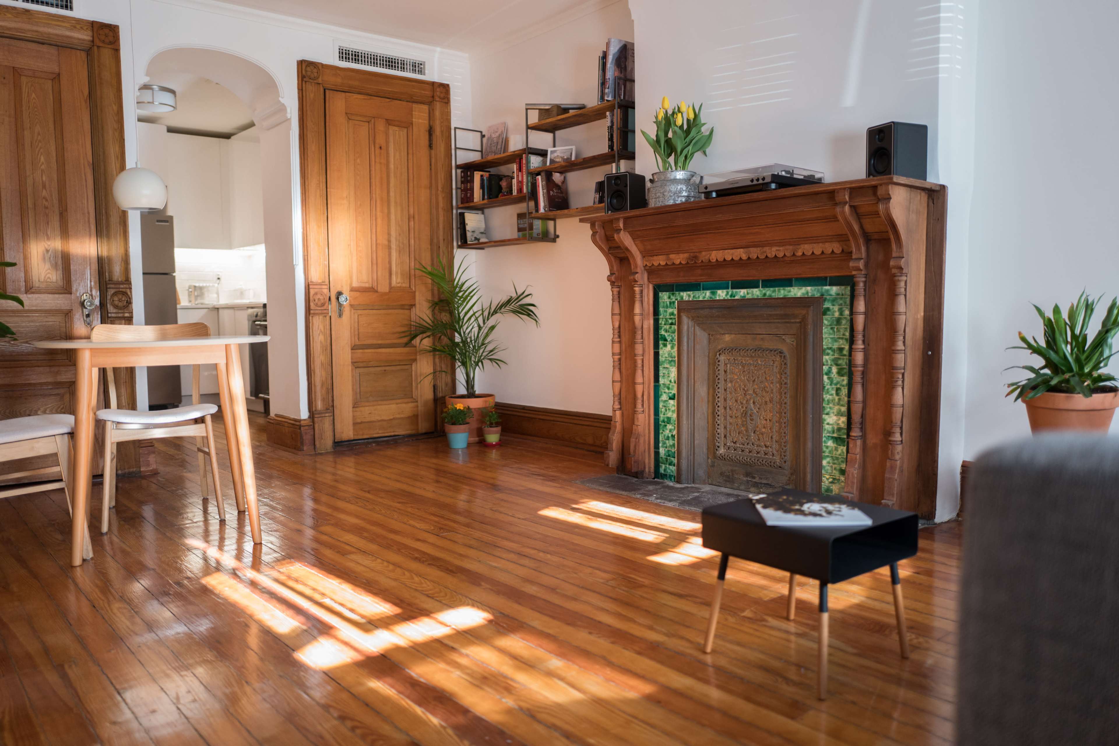 A bright room with wooden floors features a vintage fireplace, plants, and a dining table with chairs.