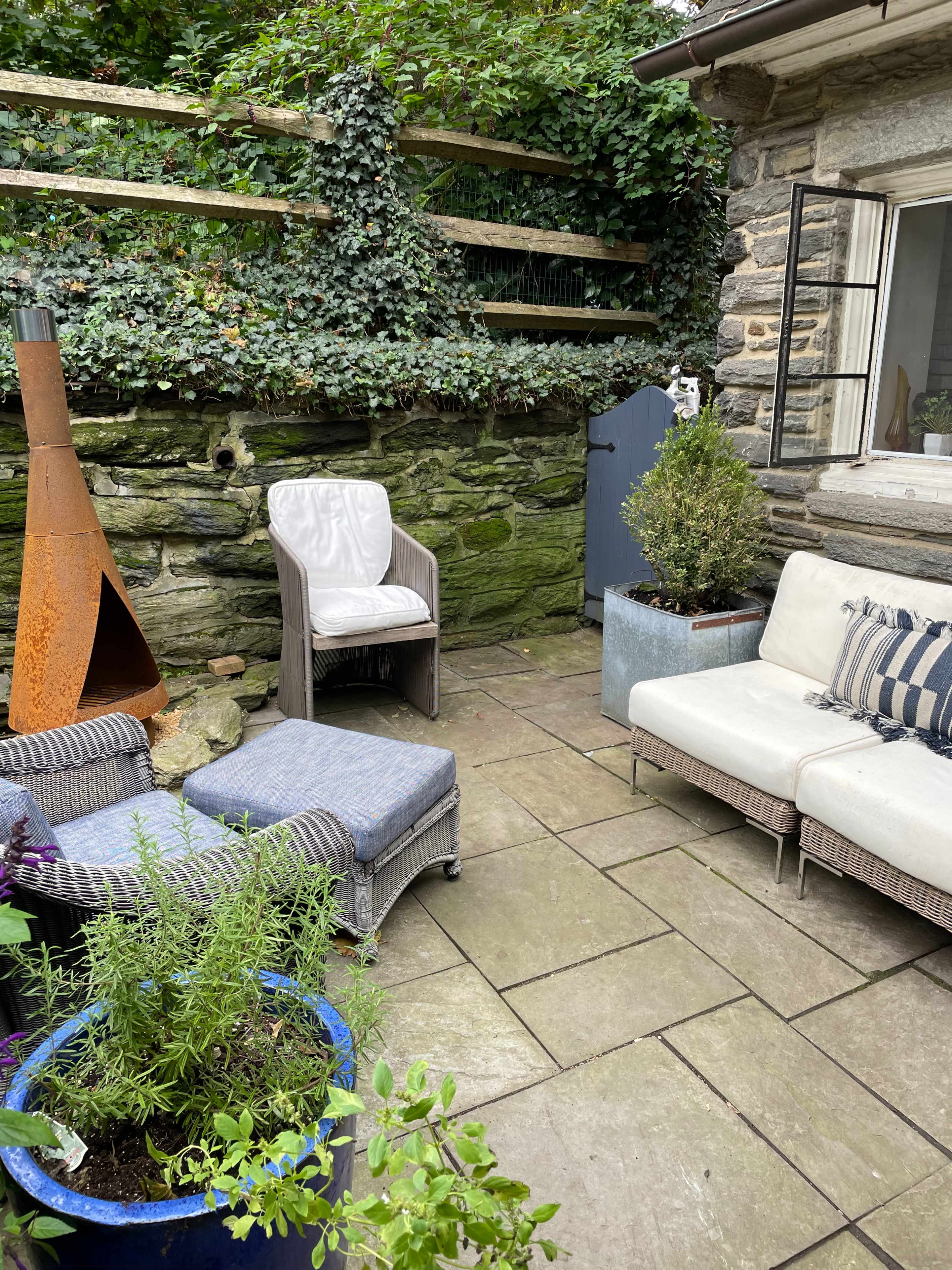 The image shows a small outdoor seating area with a mix of armchairs, a small table, and potted plants against a stone wall covered in ivy.