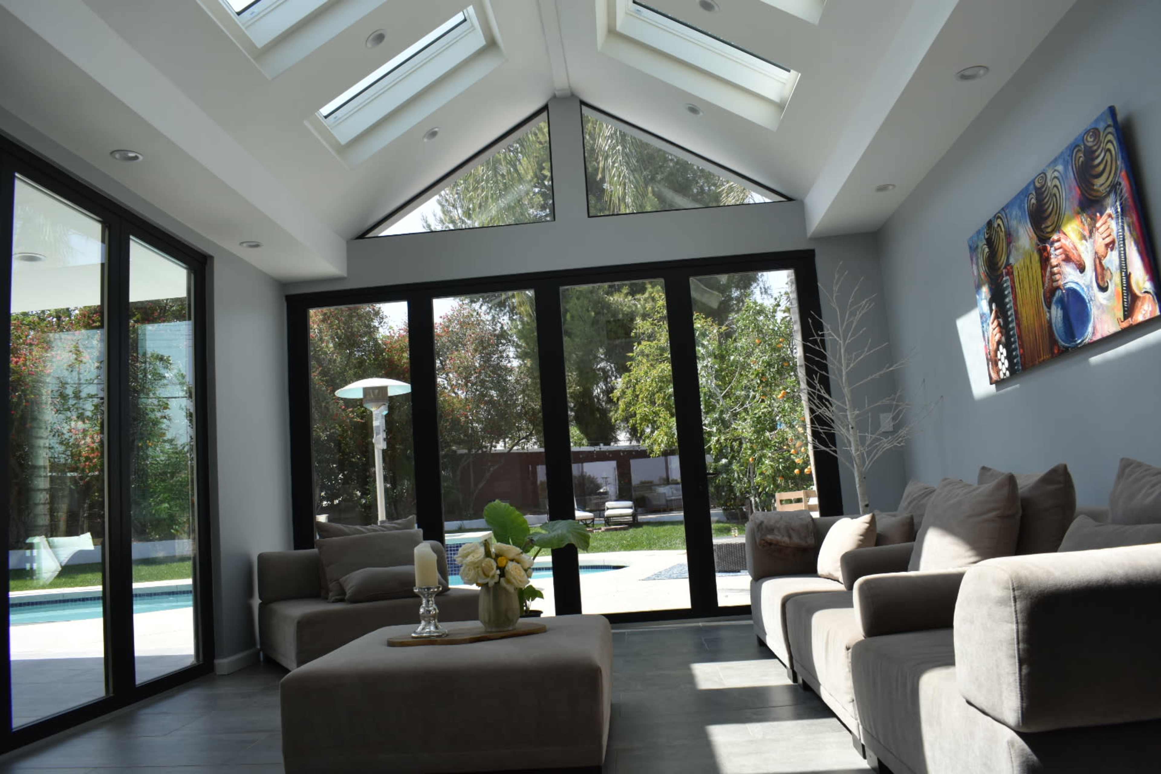 A modern living room features large glass windows, a neutral-colored sofa set, and artwork on the wall, with a view of an outdoor pool area.