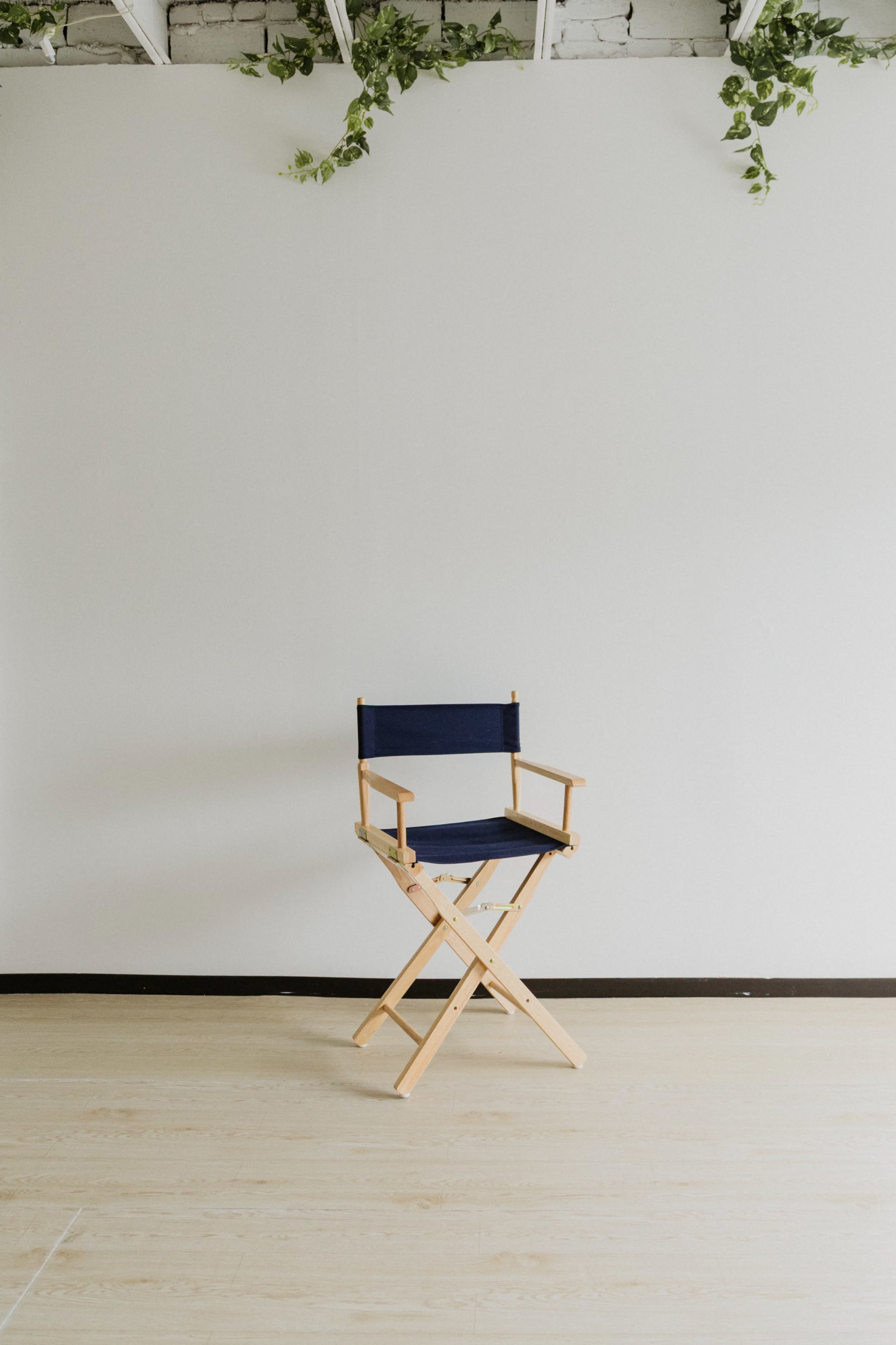 A wooden director's chair with a navy blue canvas seat is positioned on a light wood floor in front of a plain white wall.