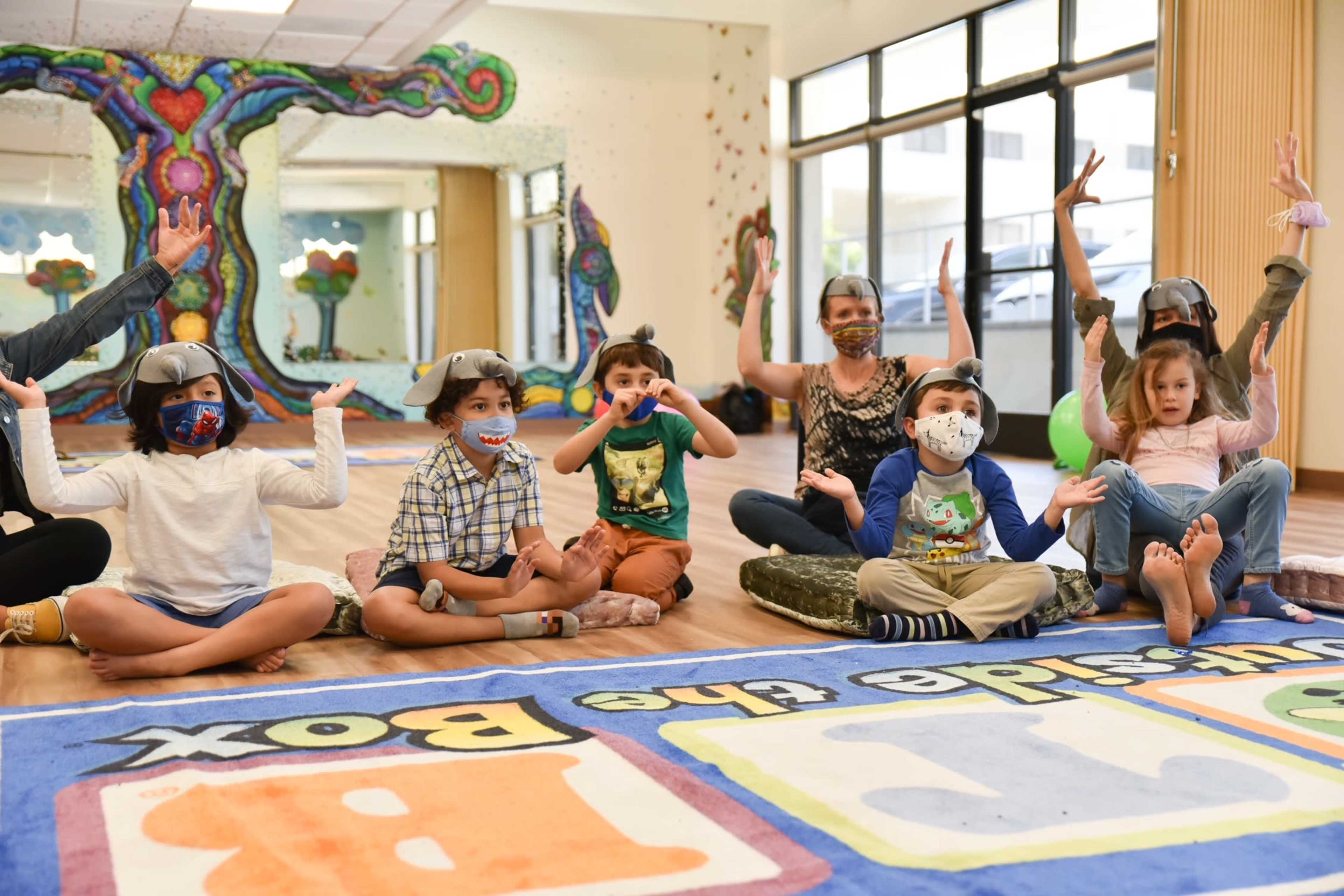 A group of children and adults sit on the floor of a colorful room, wearing masks and engaging in an interactive activity.
