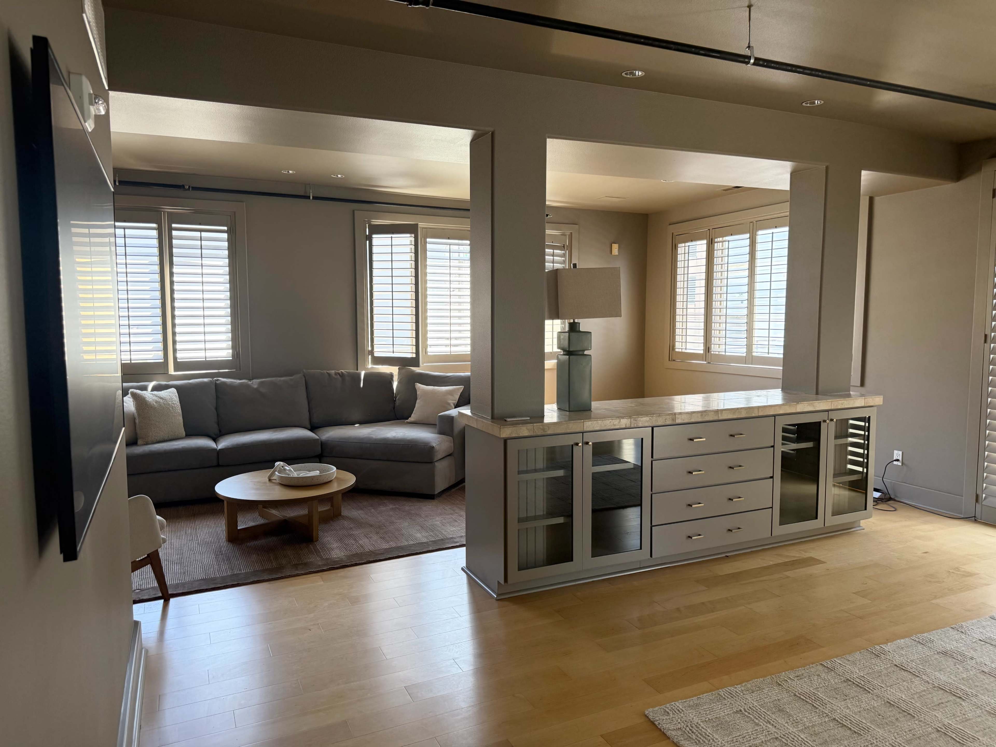 The image shows a modern living room with a gray sectional couch, a round coffee table, and a console cabinet against a wall, illuminated by natural light from windows with shutters.