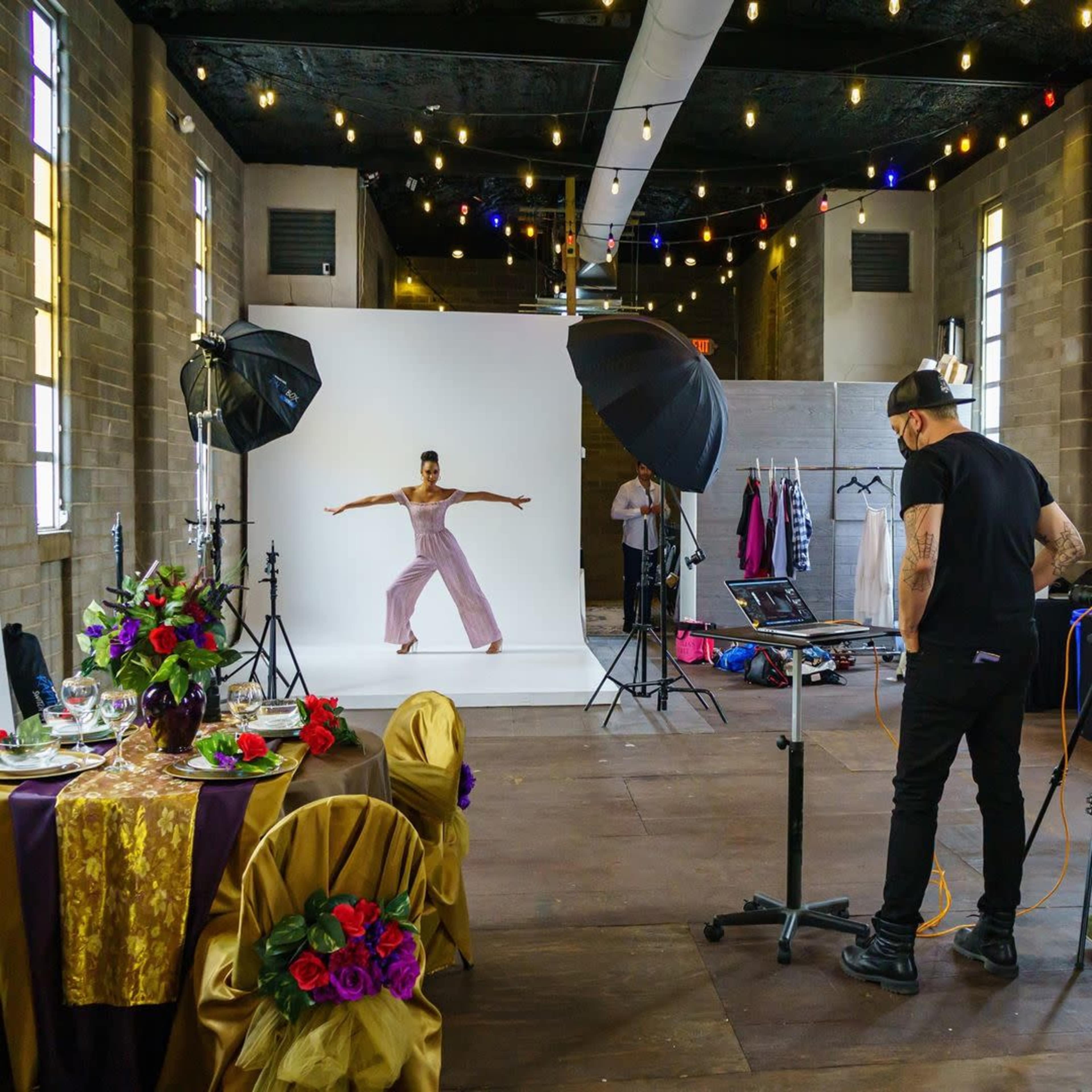 A model poses in front of a backdrop while a photographer adjusts equipment in a studio setting decorated with lights and a table set for an event.