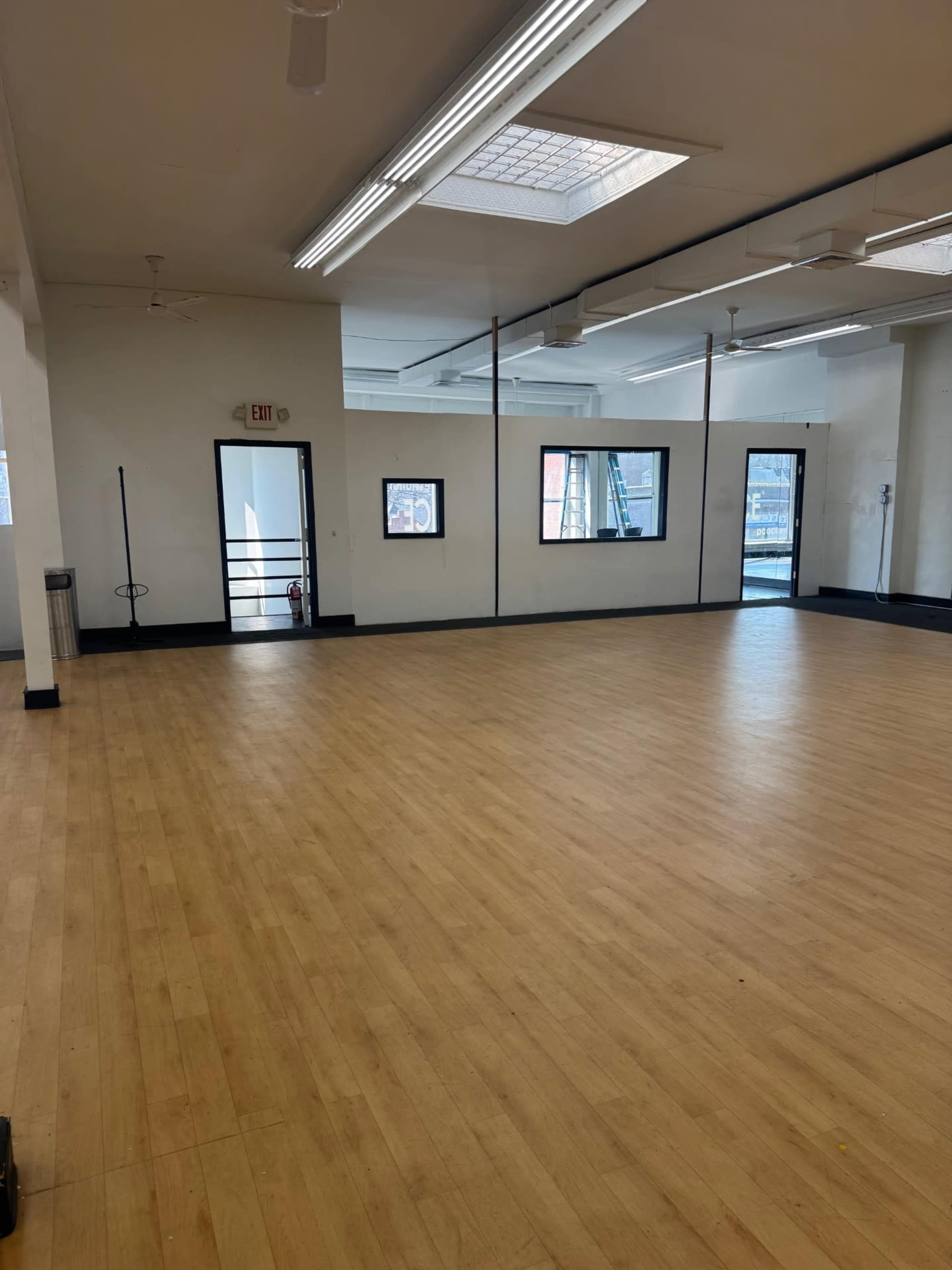 The image shows a large, empty room with wooden flooring and multiple windows reflecting overhead lights.