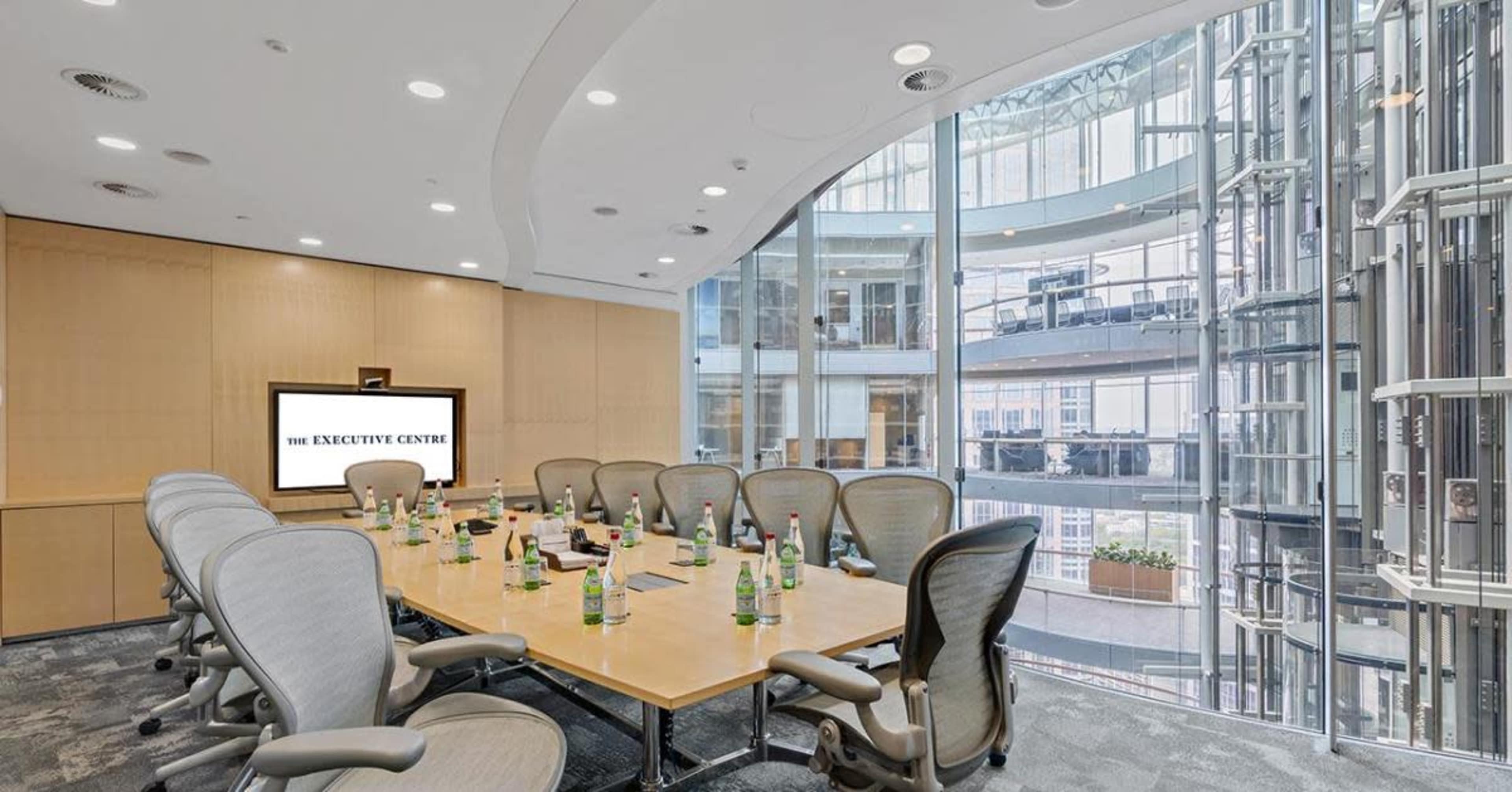 A modern conference room with a large oval table surrounded by ergonomic chairs, a presentation screen displaying "THE EXECUTIVE CENTRE," and large windows offering a view of an atrium area.