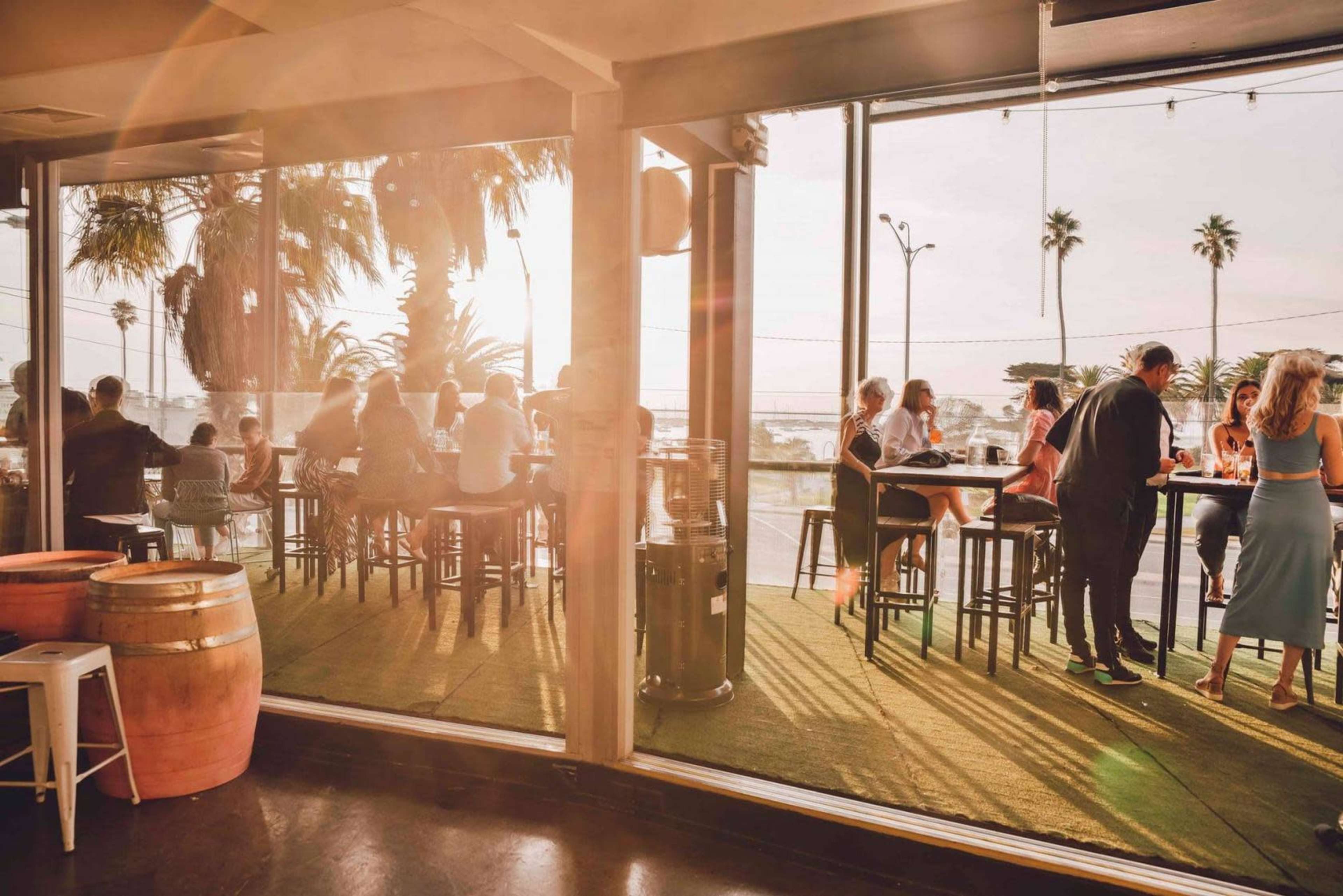 The image shows a lively restaurant scene with patrons seated at bar tables, enjoying drinks against a backdrop of palm trees and a setting sun.