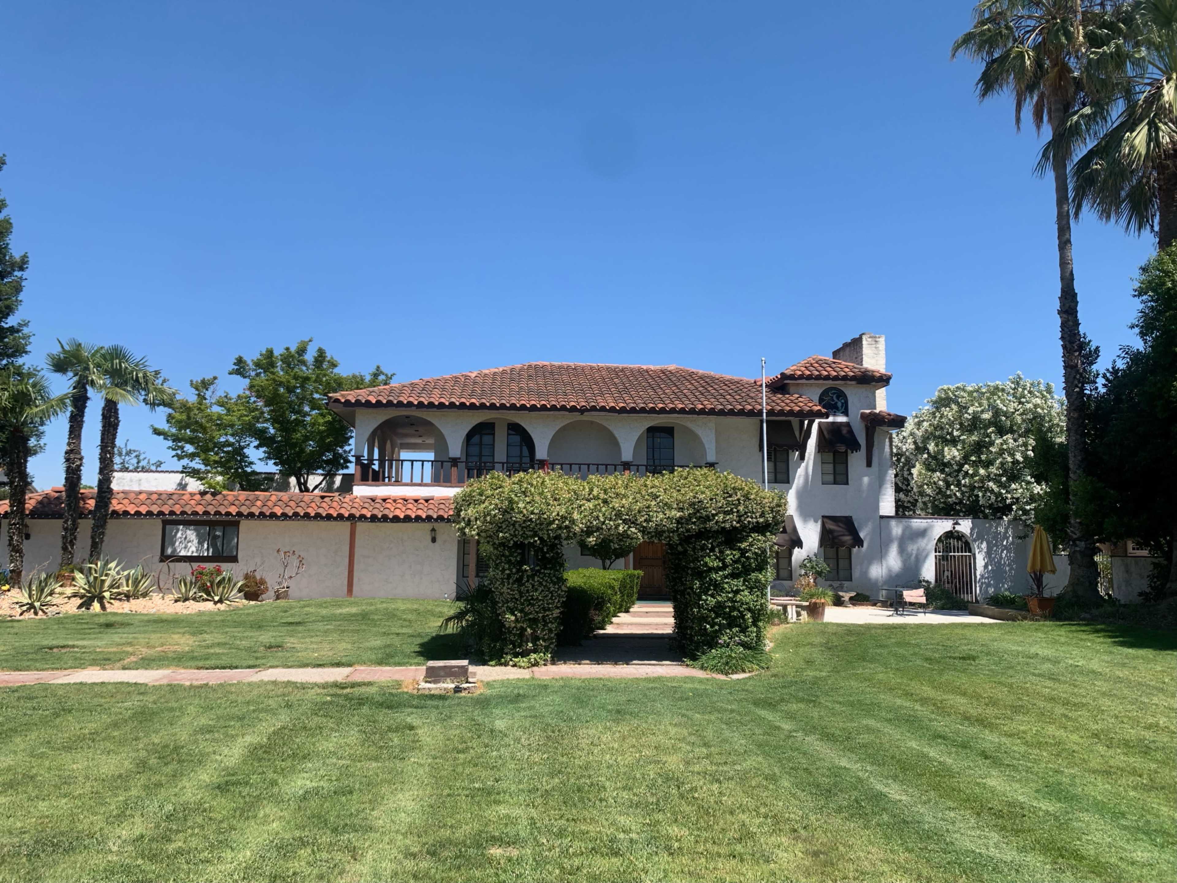 The image shows a two-story, Mediterranean-style house with a red tile roof, surrounded by well-maintained green lawns and palm trees.
