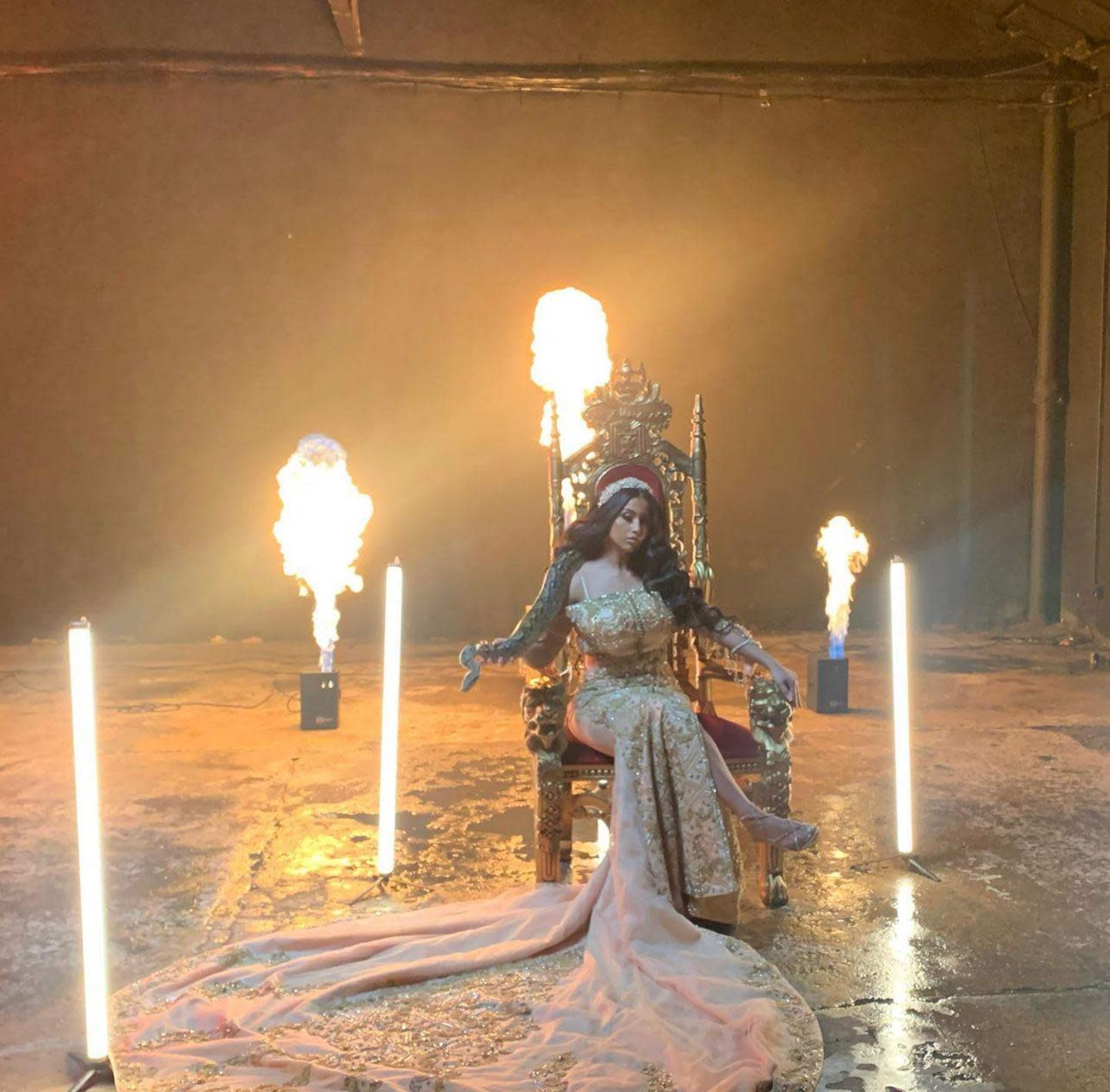 A figure in an ornate gown sits on a throne surrounded by flames and illuminated by spotlights in a dimly lit industrial space.