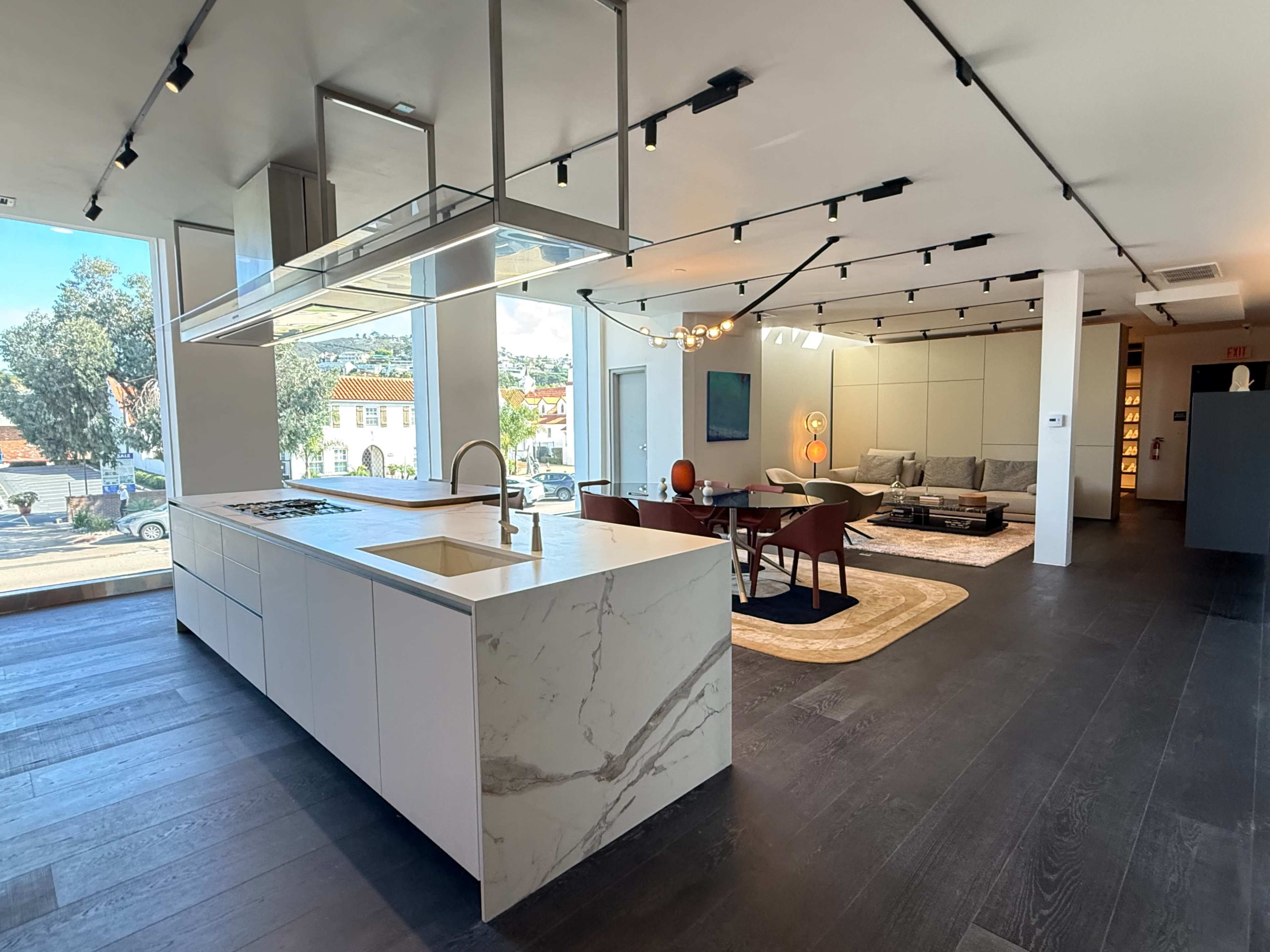 The image shows a modern, open-concept kitchen and living area with sleek cabinetry, a central island, and large windows allowing natural light to fill the space.