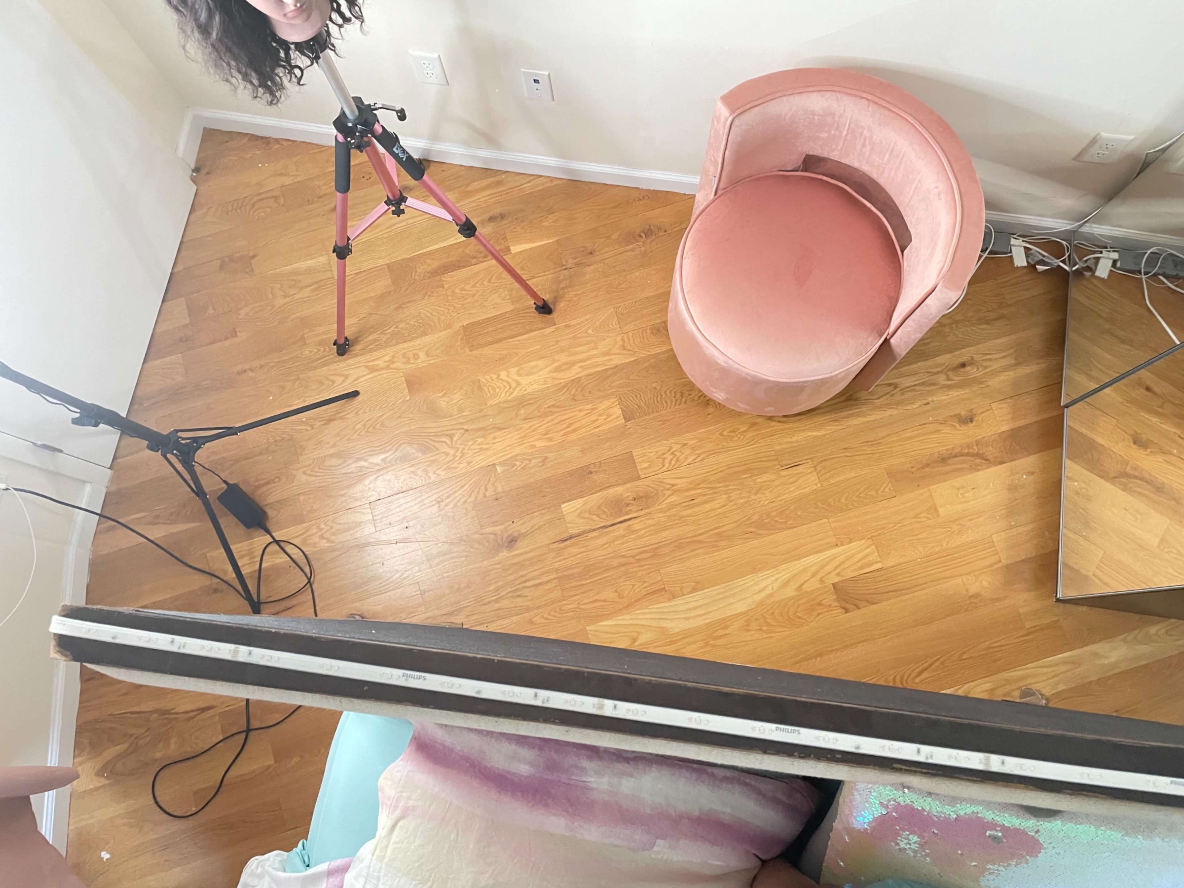 A room with a wooden floor, a pink velvet chair, a tripod with a camera, a microphone, and a large mirror.