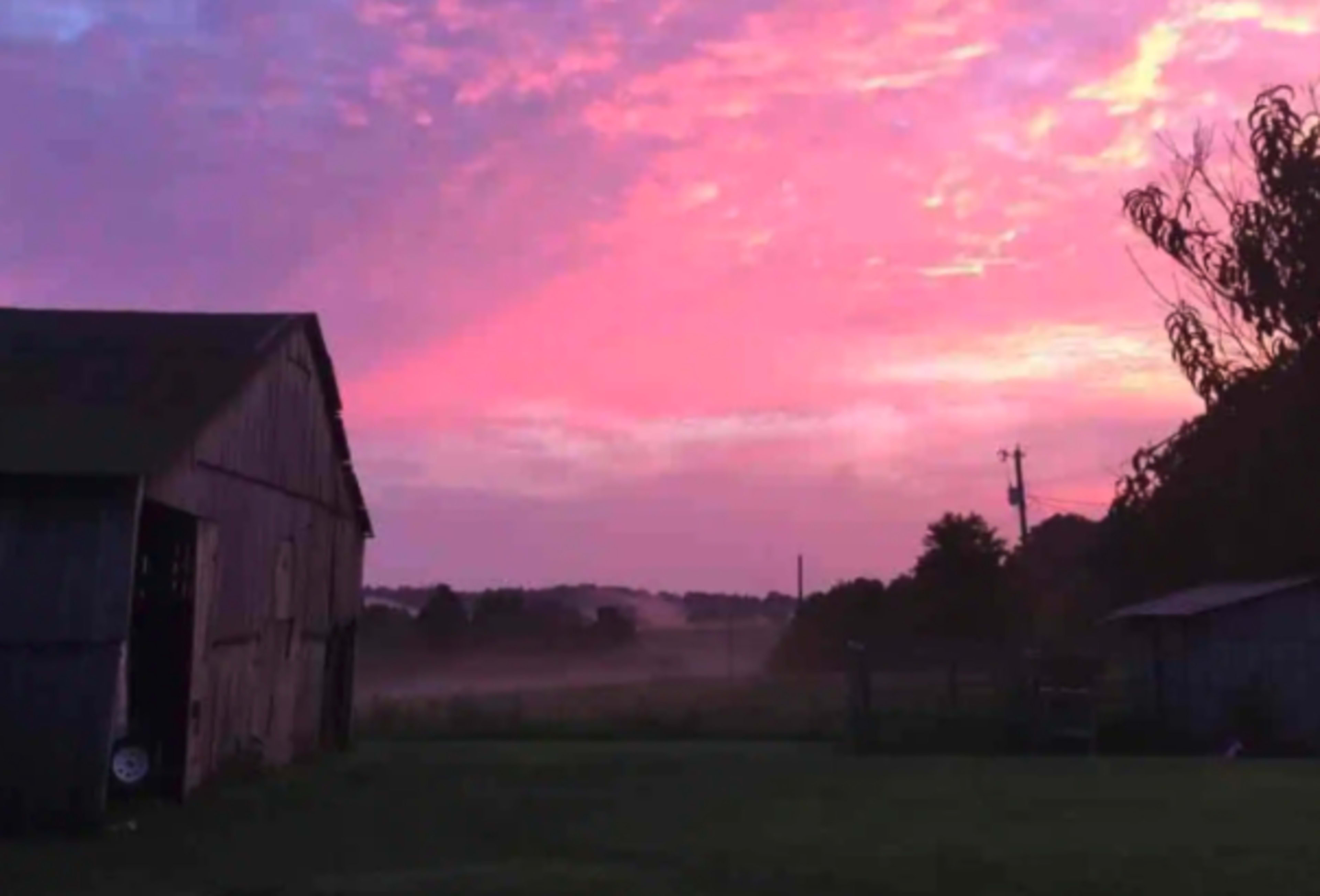 A weathered barn stands against a backdrop of a colorful sunset with mist rising in the distance.