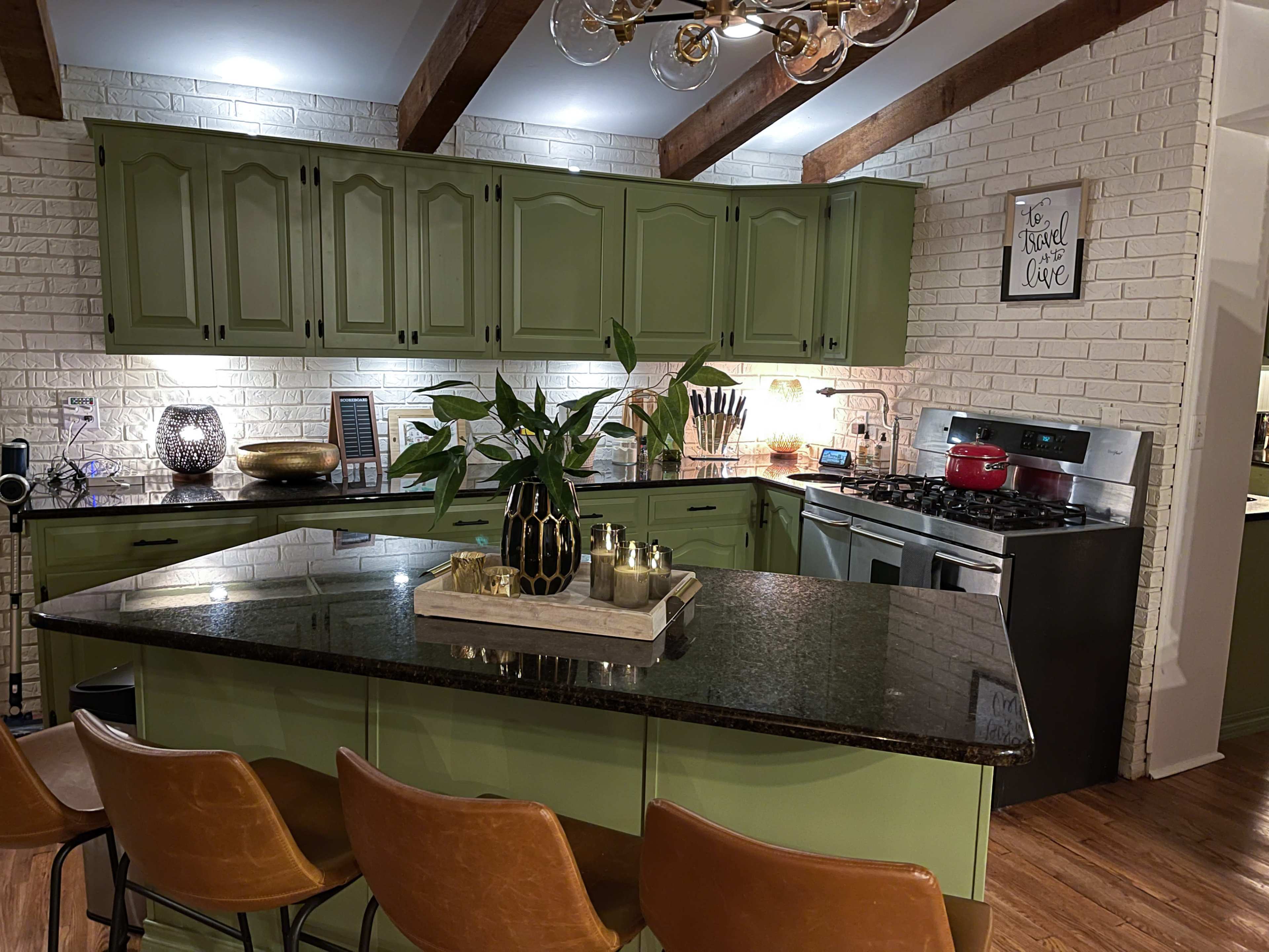 A spacious kitchen with green cabinetry, a central island topped with decorative items, and a stove against a white brick wall.