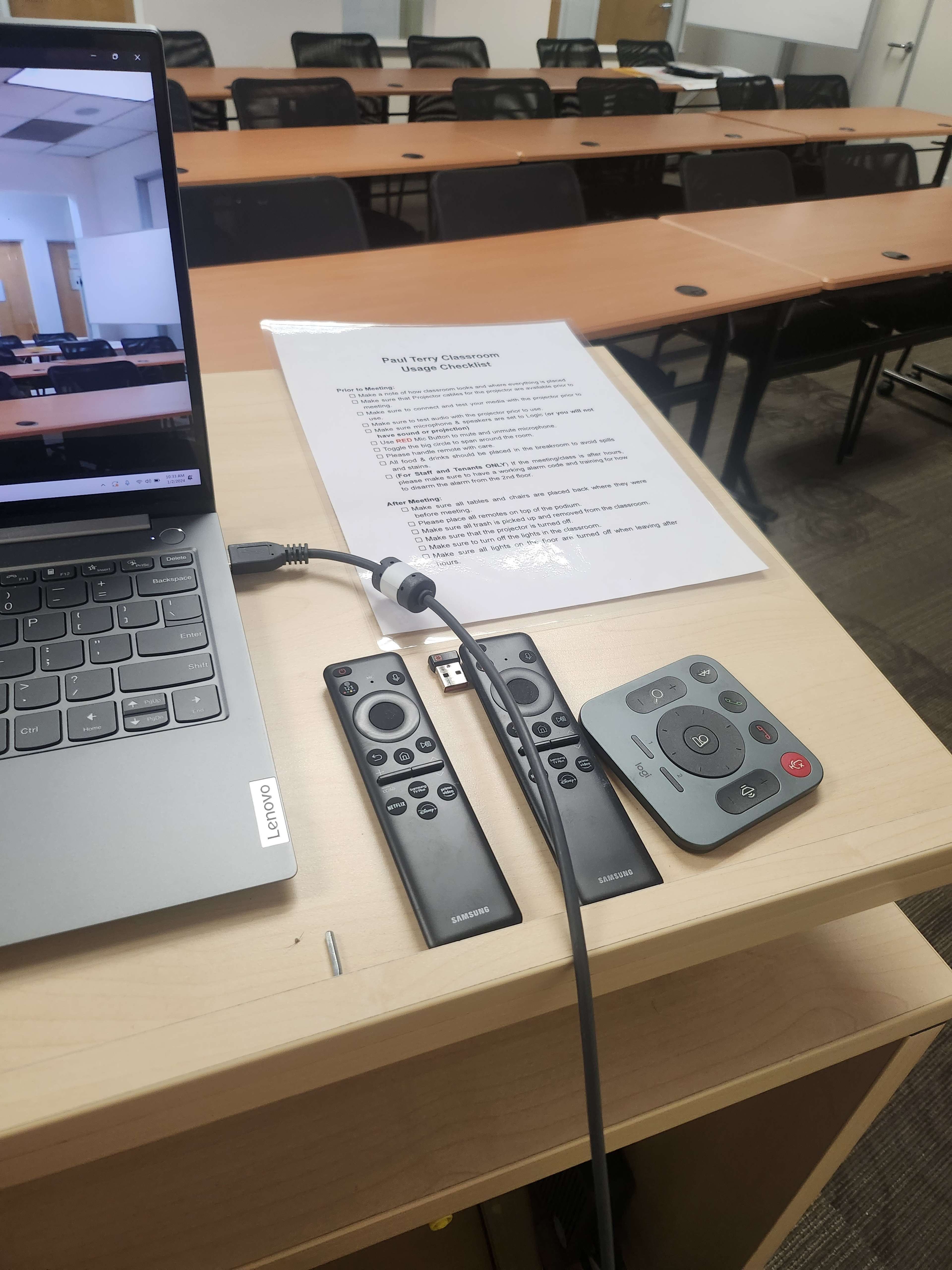 A laptop connected to two remote controls and a USB adapter is placed on a table in a classroom set up for a meeting or presentation.