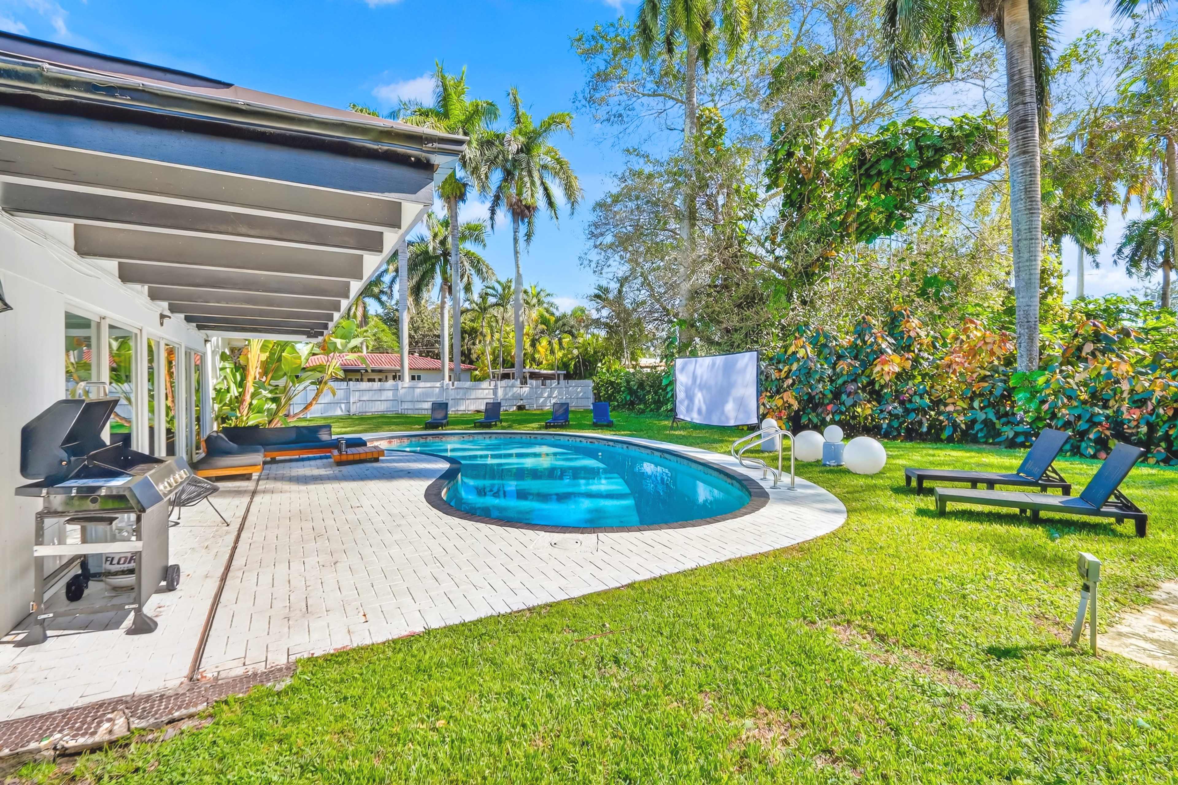 The image shows a backyard with a curved swimming pool surrounded by palm trees, lounge chairs, and a patio area with a grill.