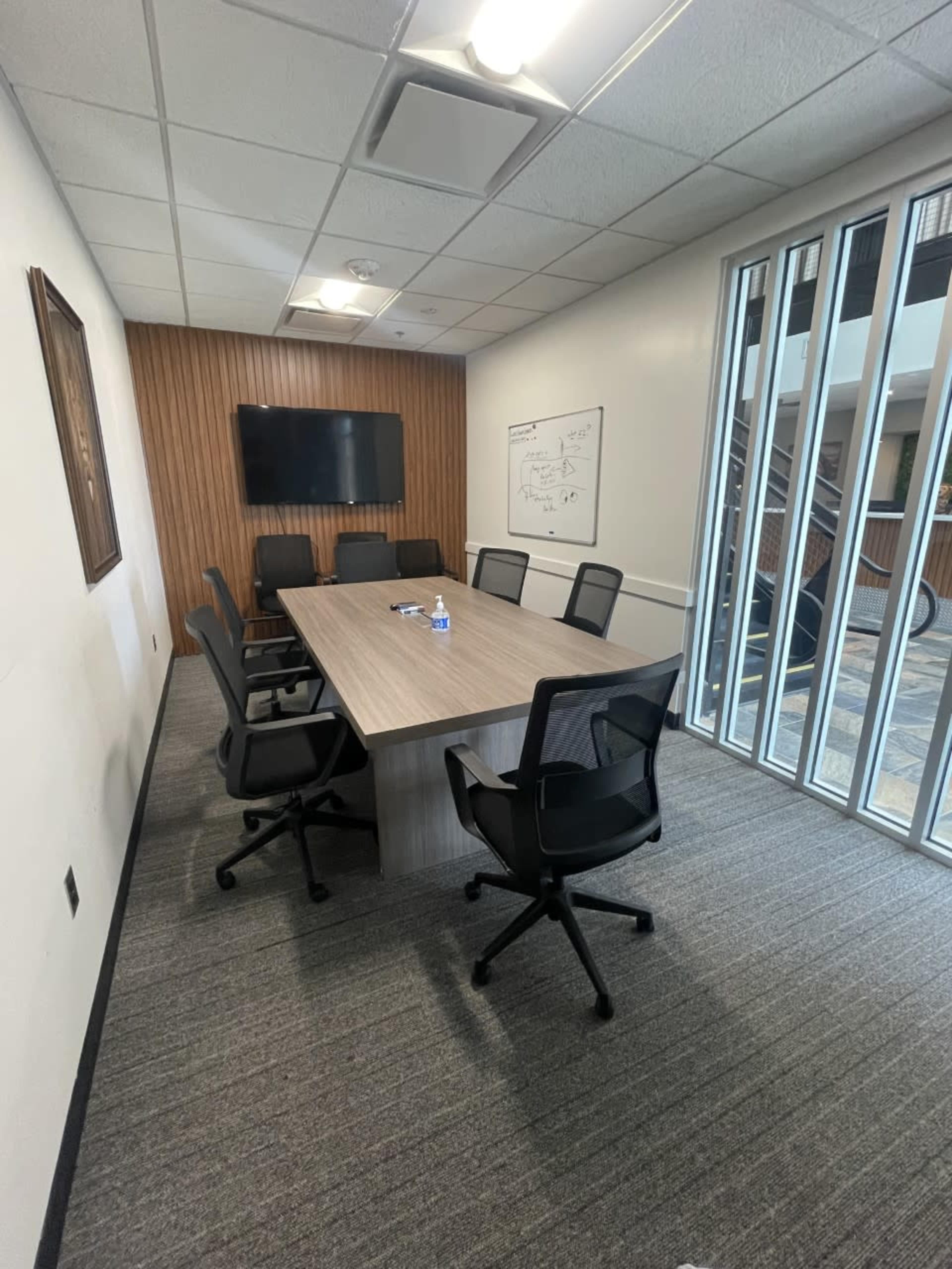 A conference room features a long wooden table surrounded by six black chairs, with a television mounted on the wall and large windows allowing light from an adjacent hallway.