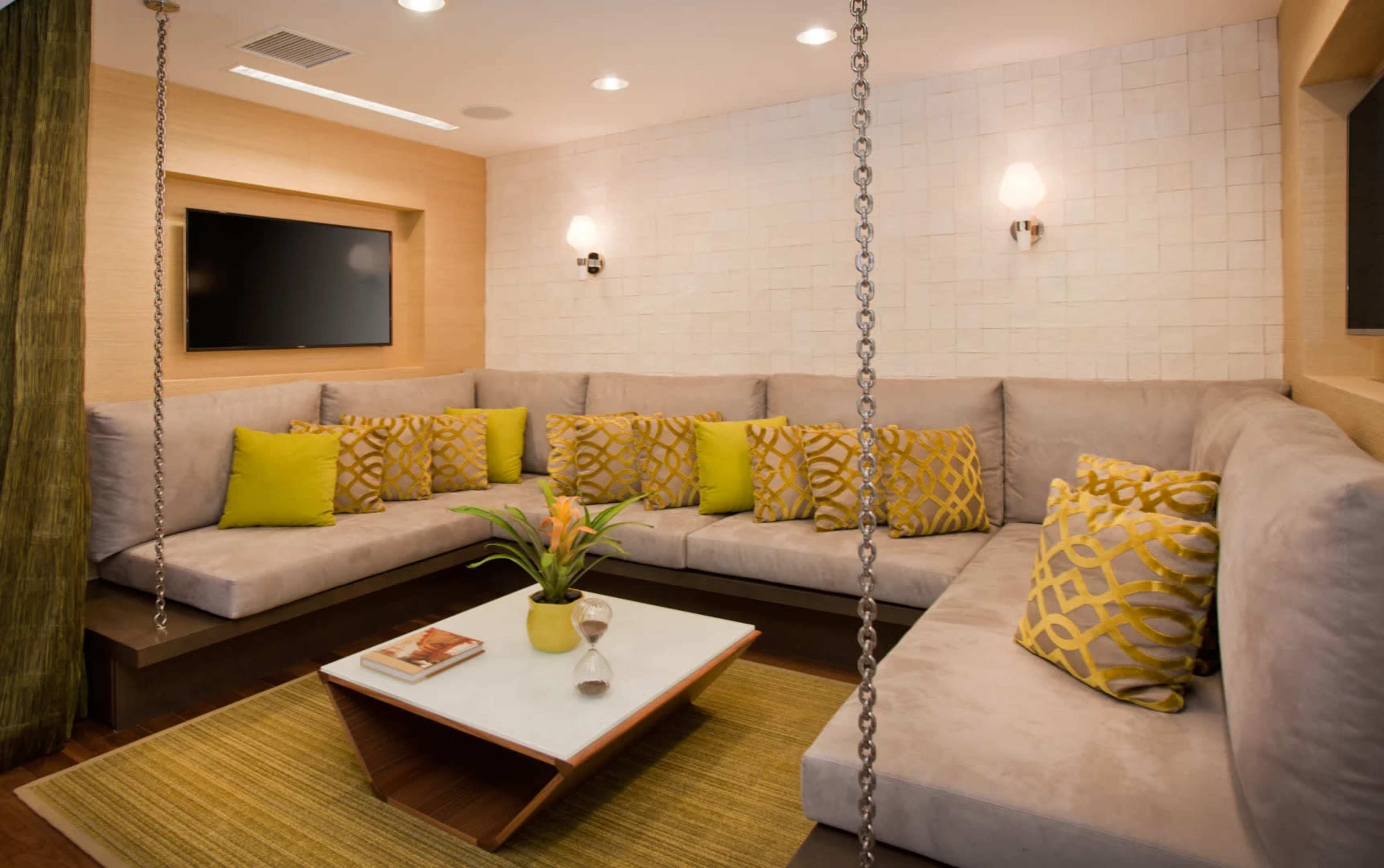 A modern living room featuring a large gray sectional sofa with yellow accent pillows, a coffee table, and a decorative plant, all arranged in a well-lit space with a television mounted on the wall.