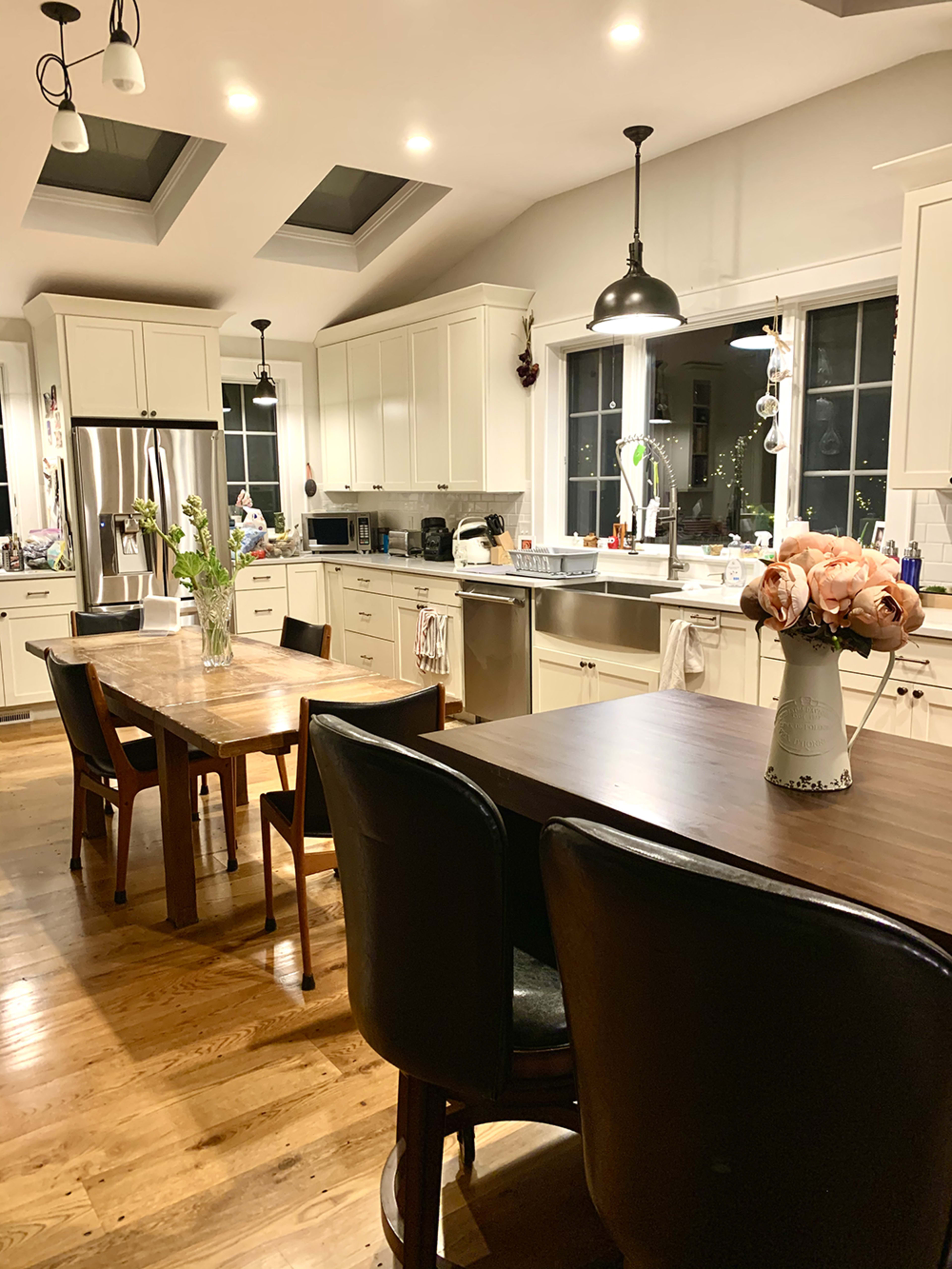 A spacious kitchen features white cabinetry, a wooden dining table, and two black chairs, illuminated by pendant lights and skylights.