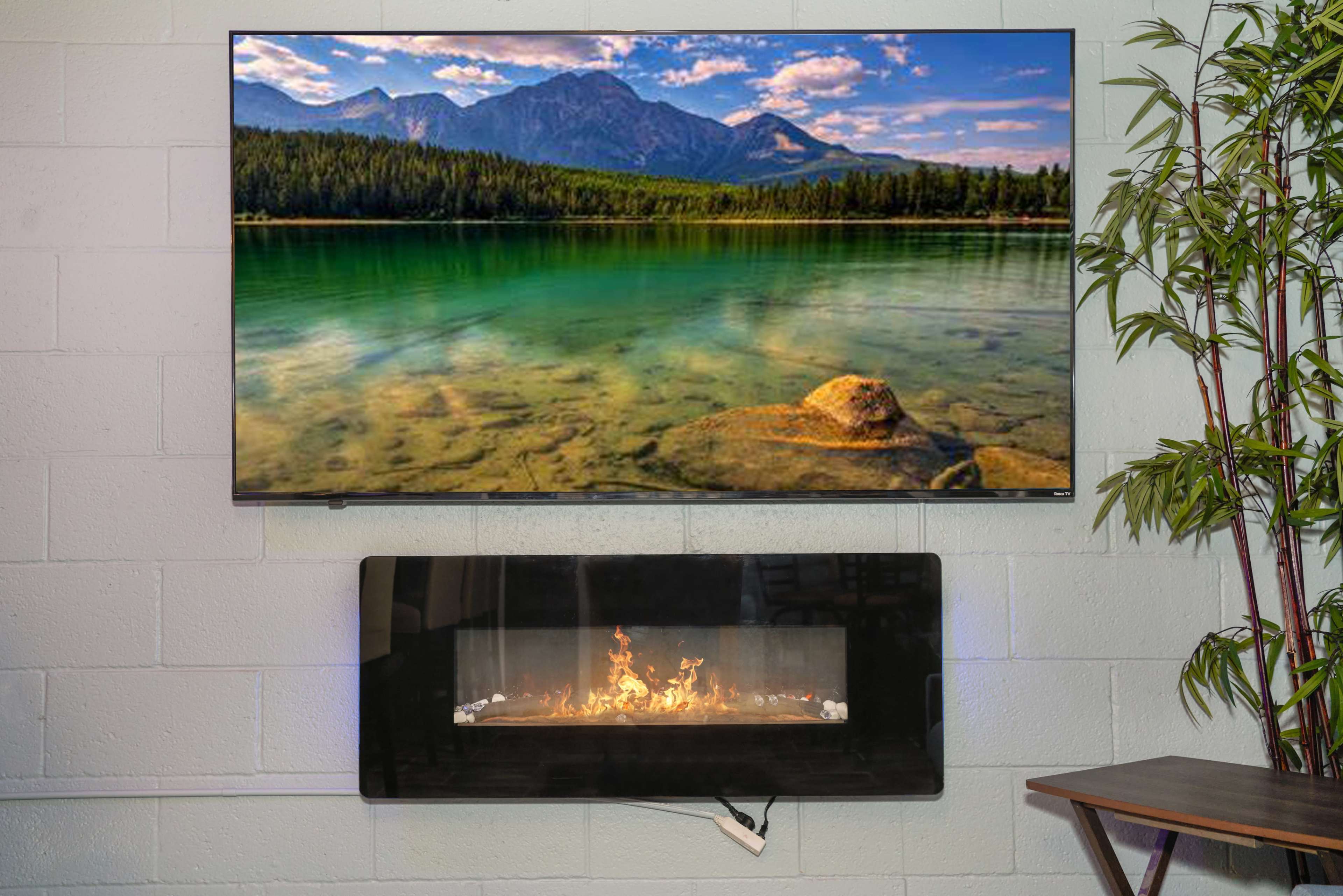 A television displaying a scenic mountain and lake landscape is mounted above a modern, wall-mounted electric fireplace.