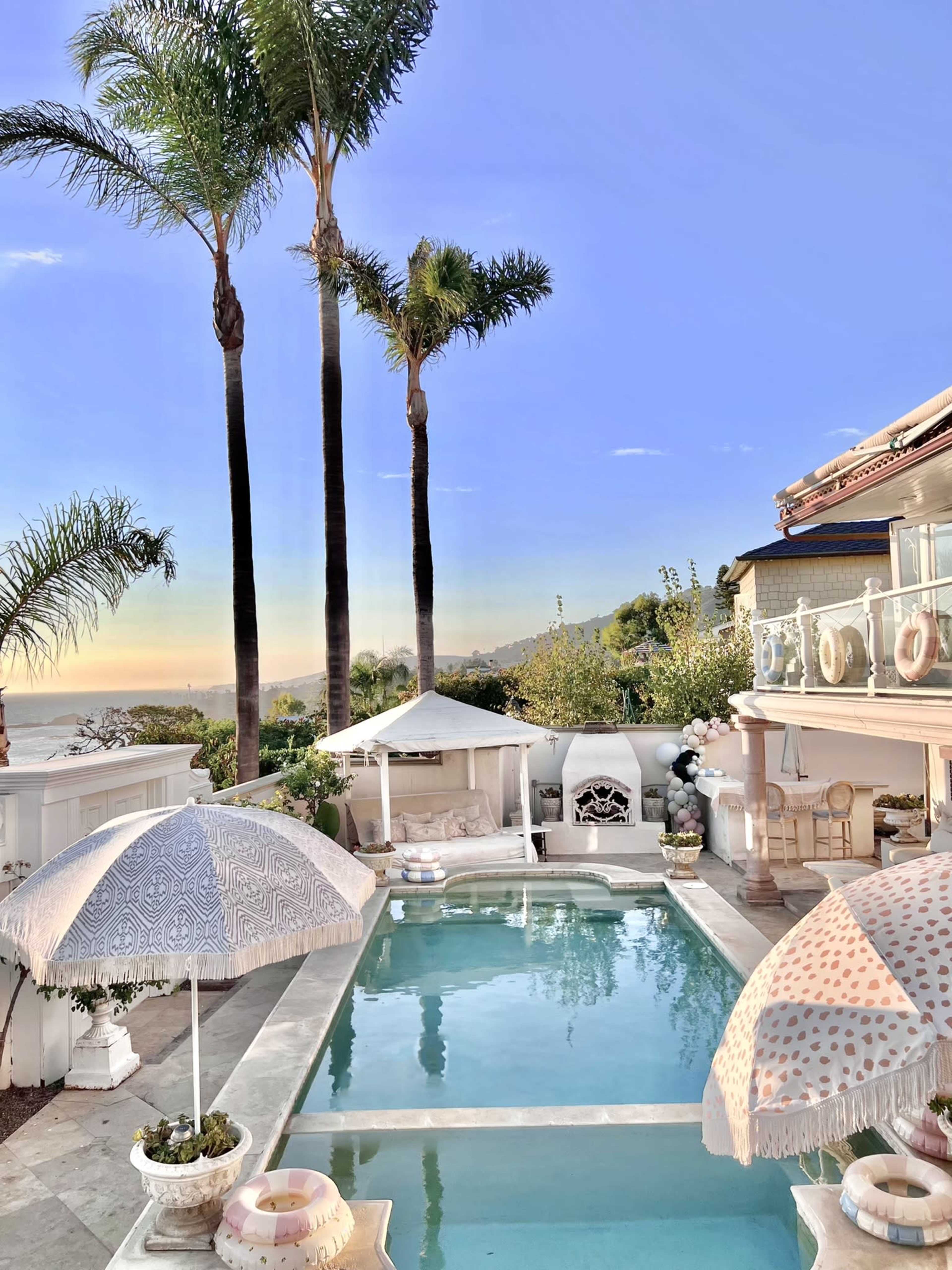 The image shows a backyard pool area with palm trees, lounge chairs, and umbrellas, overlooking a scenic view at sunset.