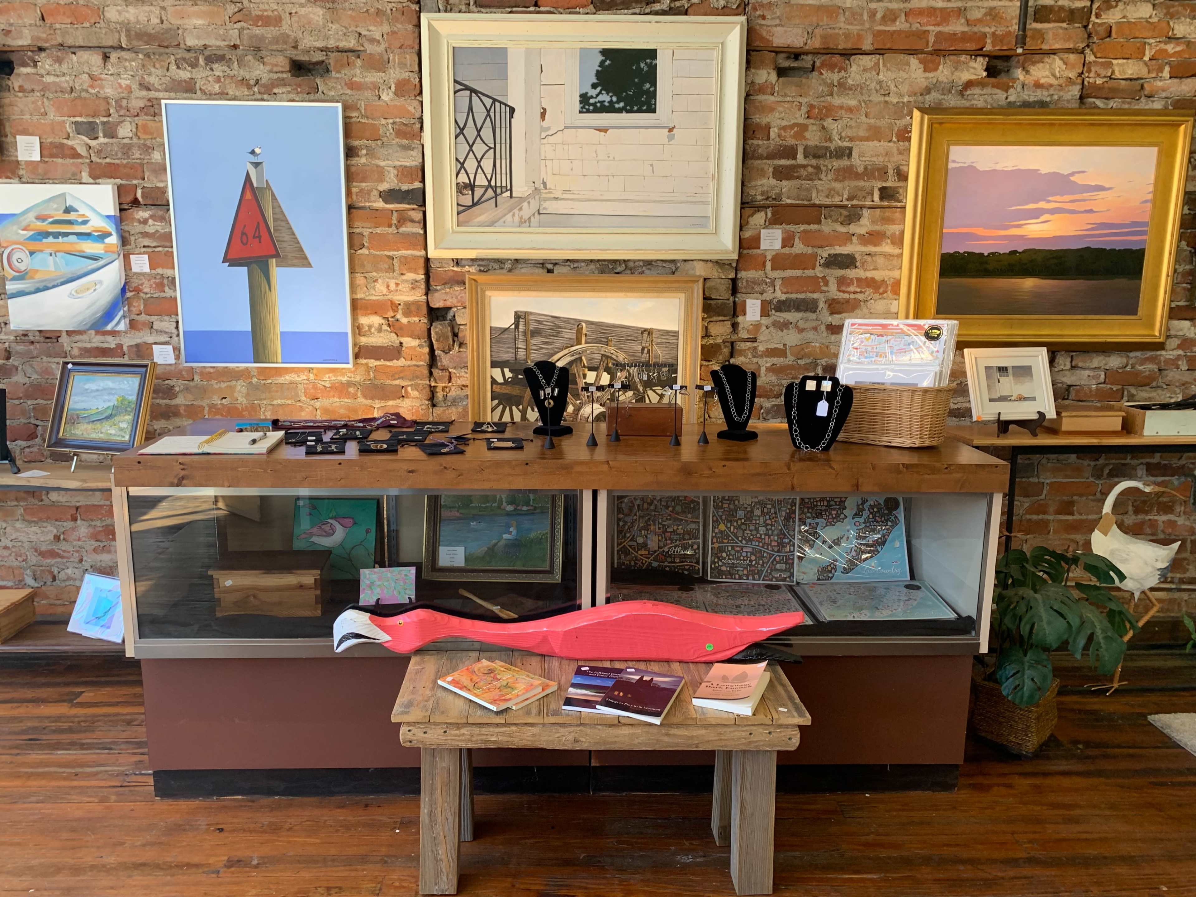 The image features an art gallery display with a wooden counter showcasing various artworks, jewelry, and decorative items against a backdrop of exposed brick walls.