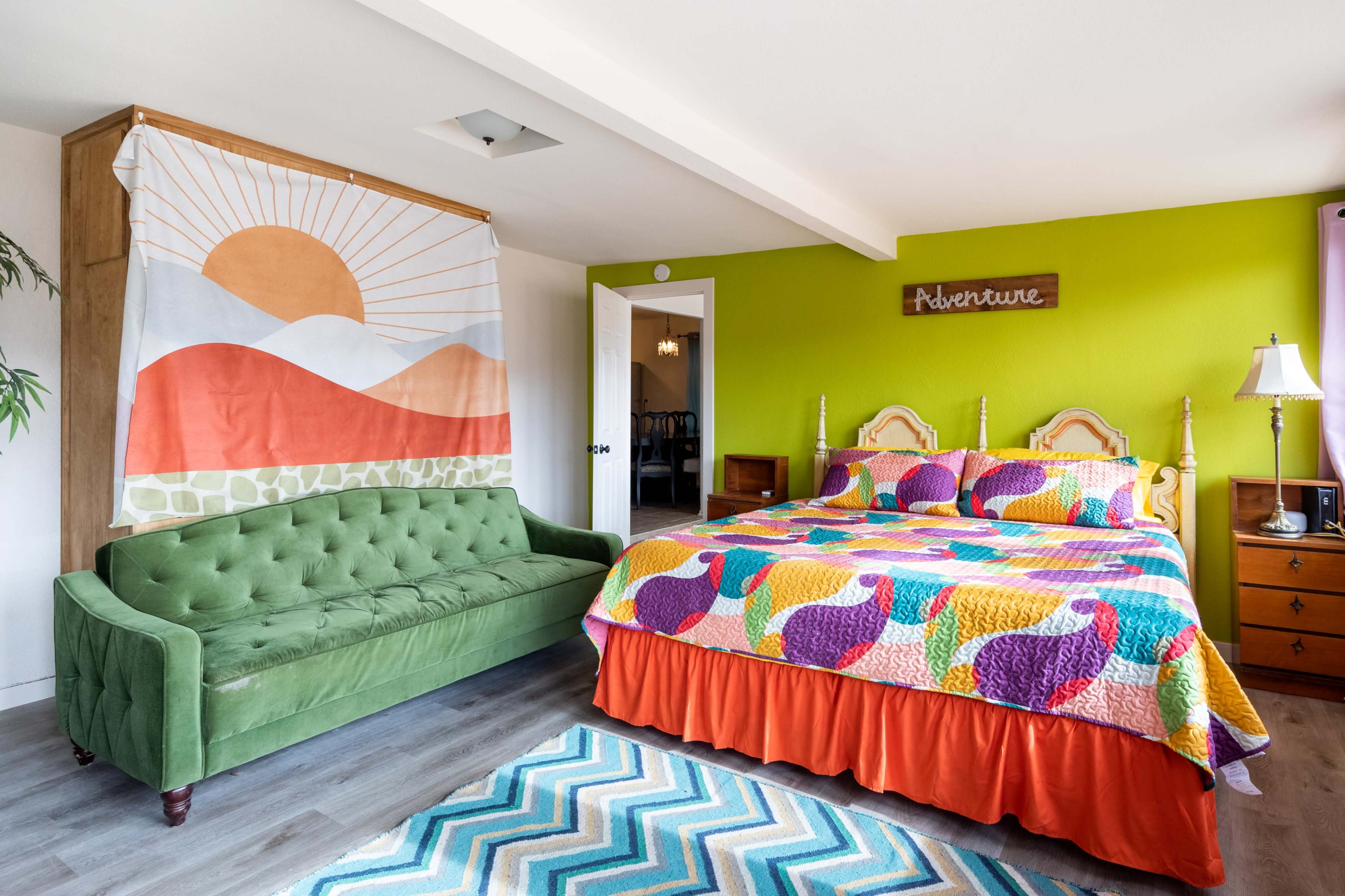 The room features a green velvet sofa, a brightly colored quilted bedspread, and a vibrant accent wall.