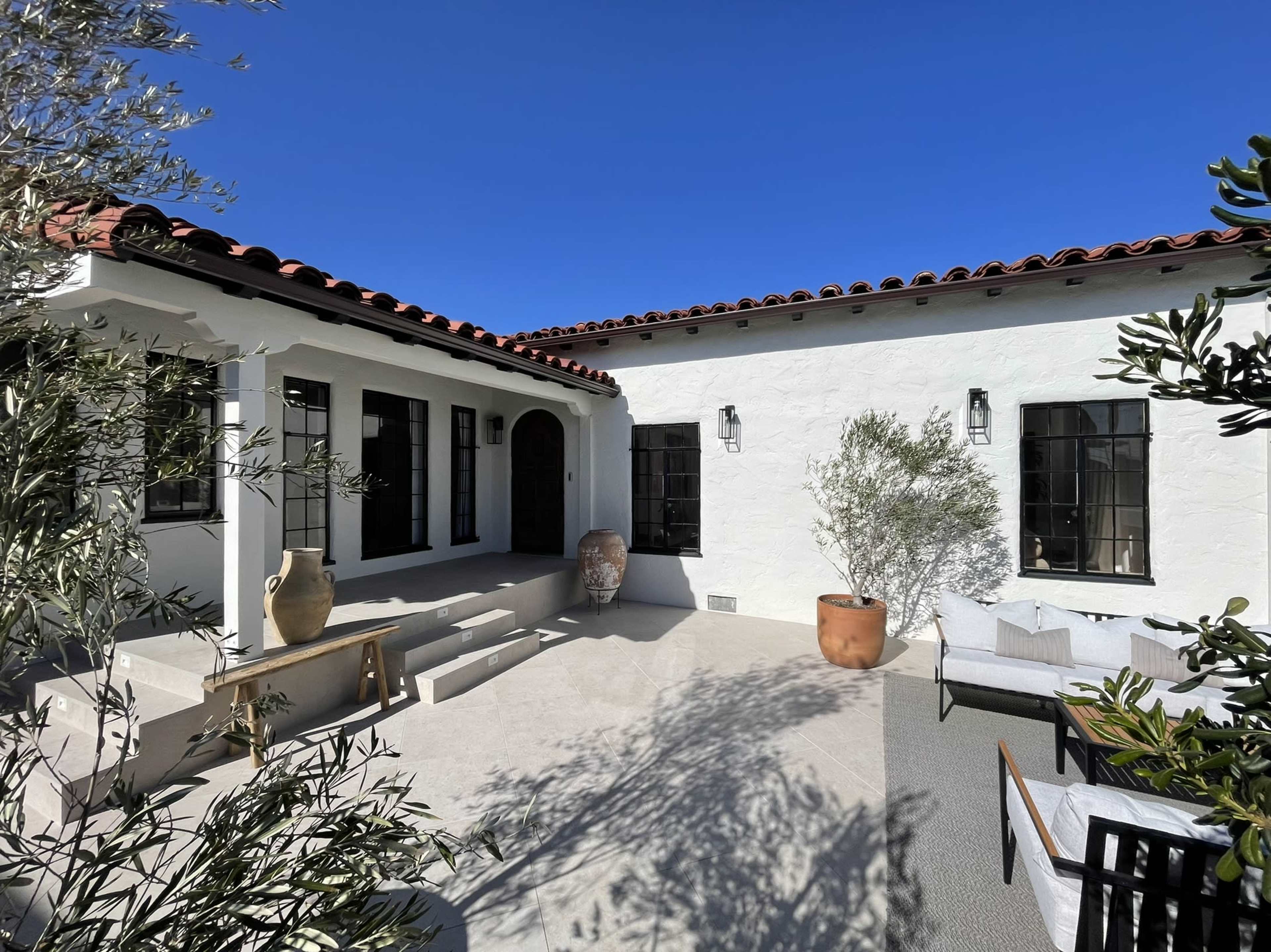 A modern courtyard features white walls, a red-tiled roof, large windows, and potted plants.