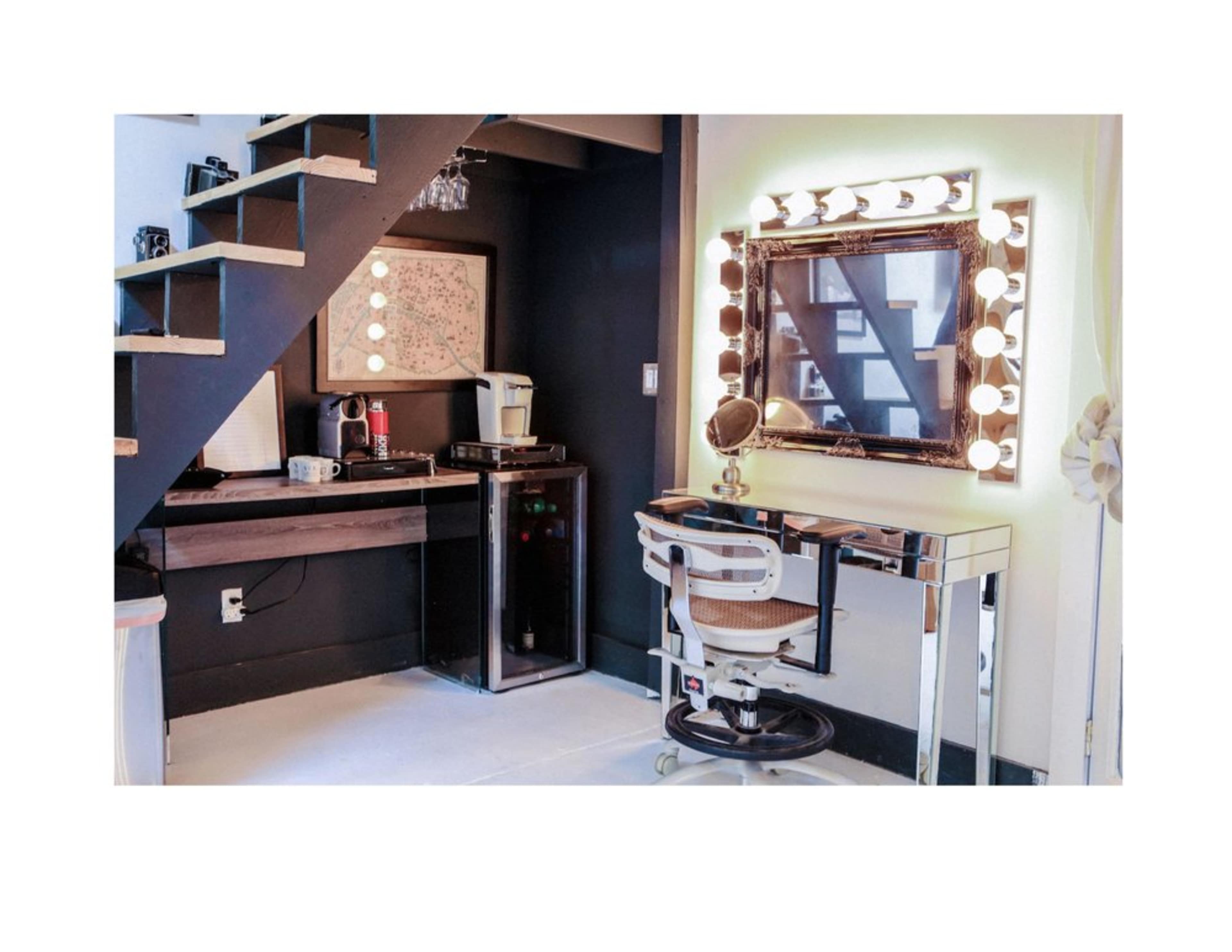 The image shows a stylish vanity area with a large mirror surrounded by lights, a desk with a coffee maker, and a staircase in the background.