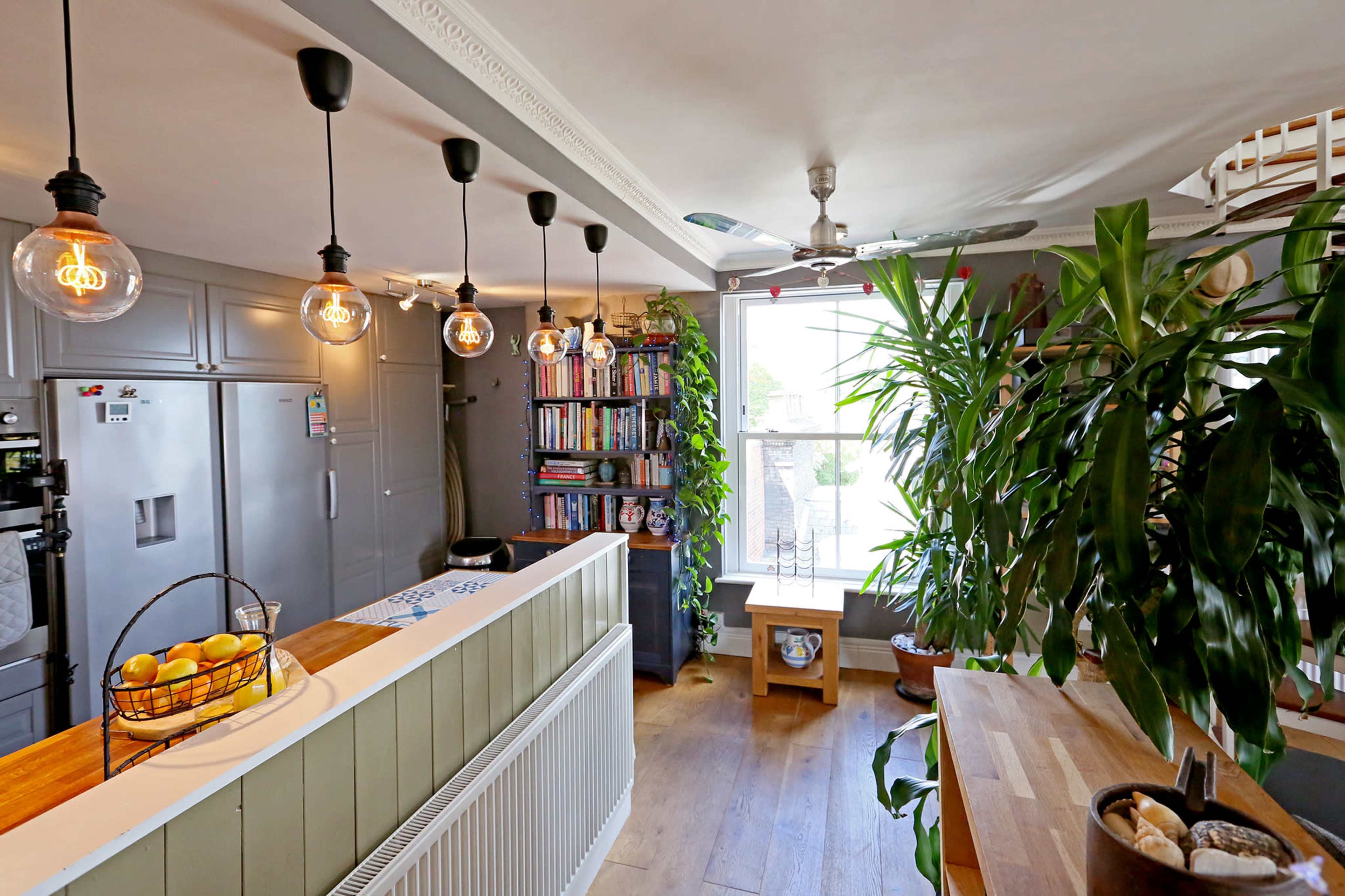 A bright, contemporary kitchen area featuring a curved countertop, hanging pendant lights, a bookshelf, and large windows with indoor plants.