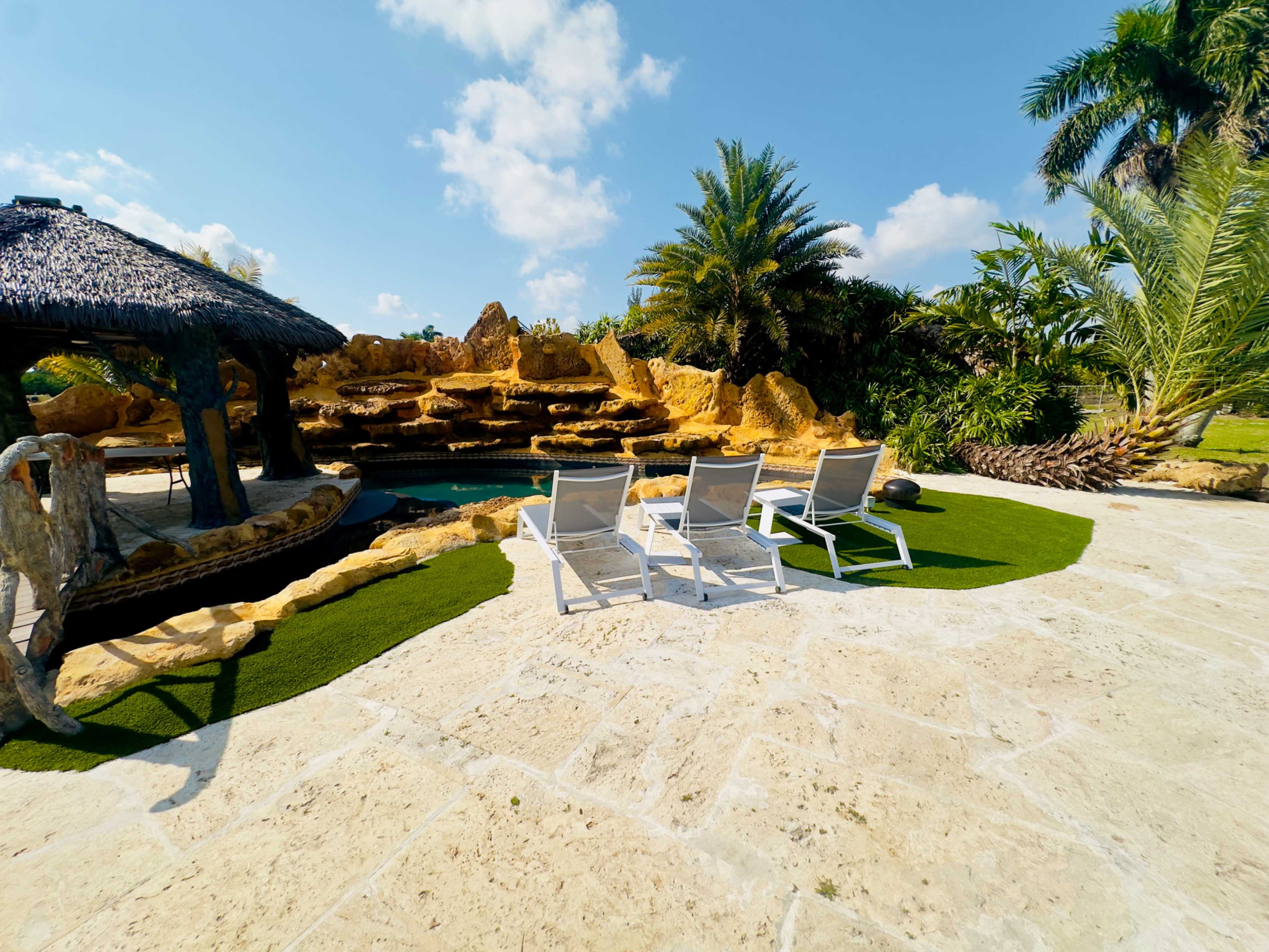 The image shows a tropical pool area featuring three lounge chairs arranged on a stone patio, surrounded by lush greenery and a waterfall.