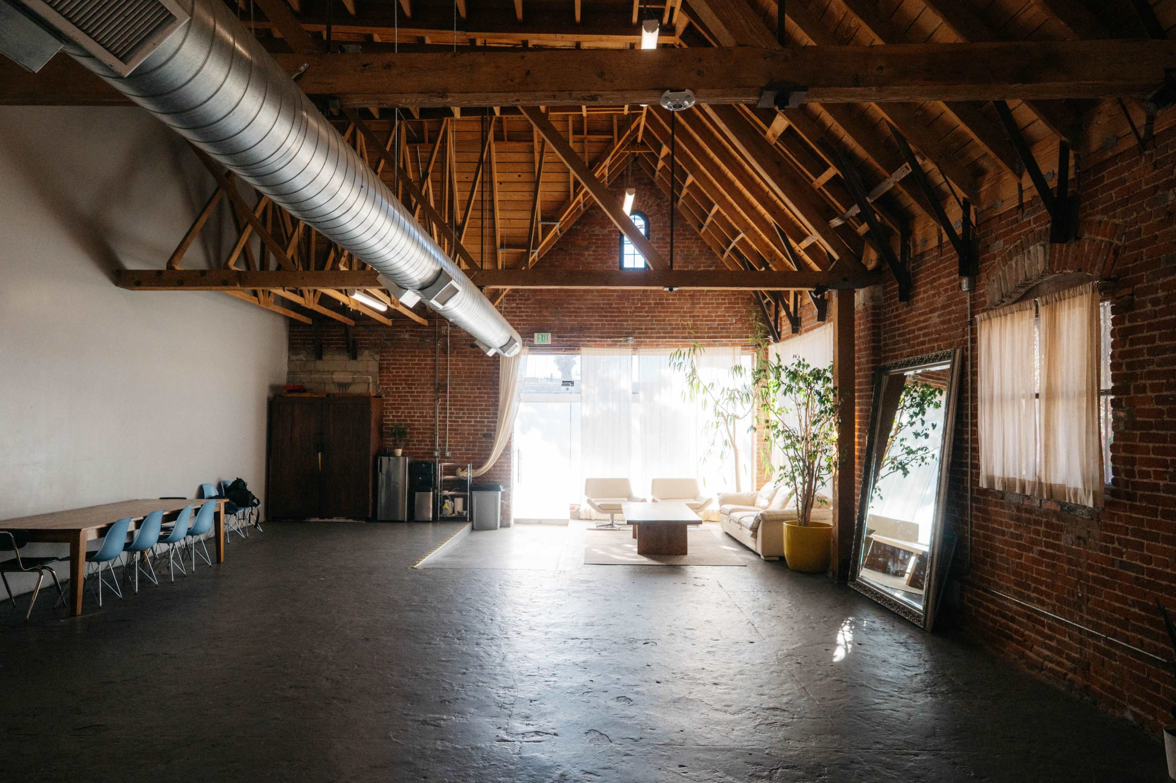 The image shows an industrial-style loft with exposed wooden beams, brick walls, and a spacious area featuring a seating arrangement, a large mirror, and plants near a window.