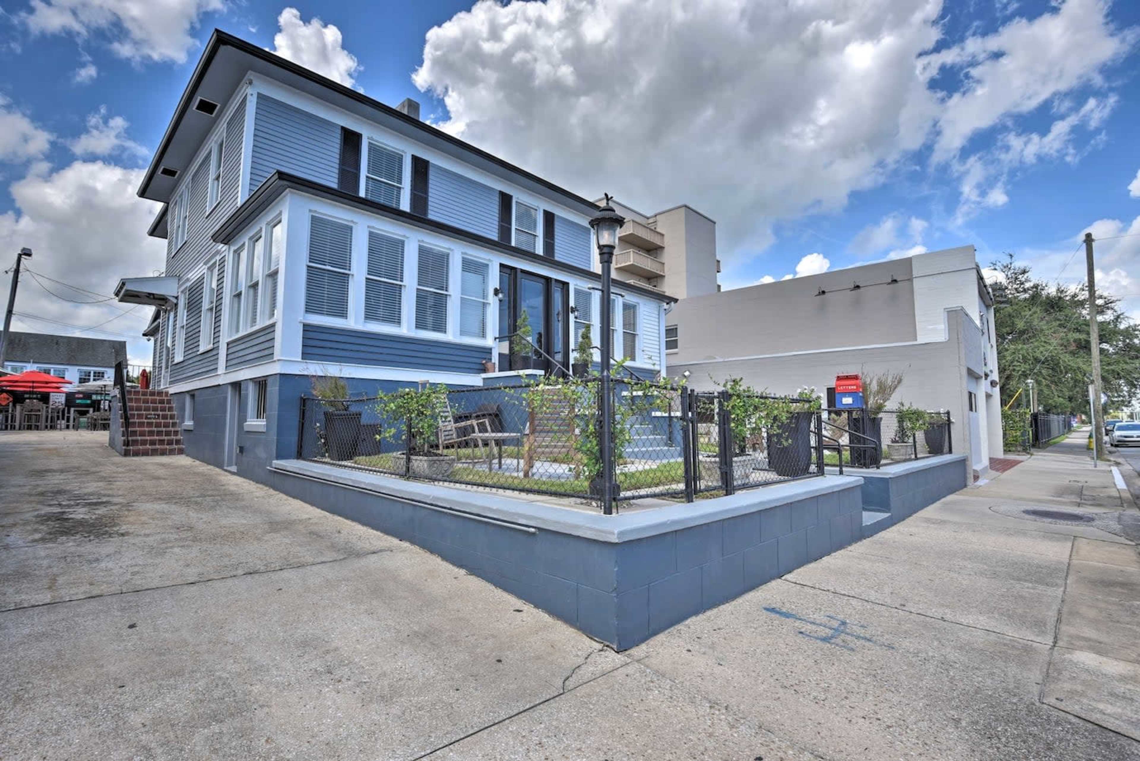 A two-story blue house with white trim, featuring a porch and surrounding greenery, sits at the intersection of a street next to a taller building.