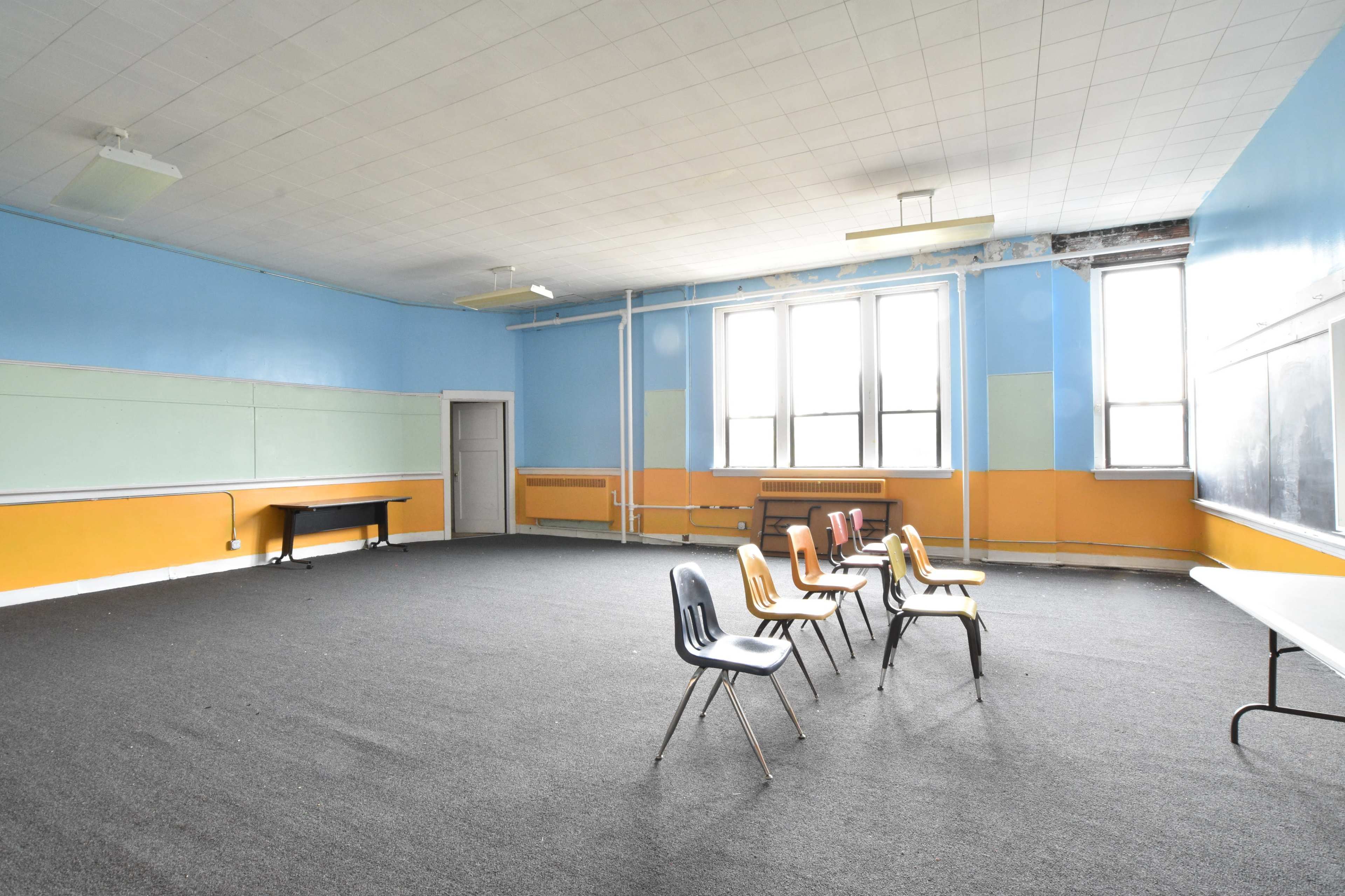 An empty classroom features a row of chairs positioned in the center, with orange and blue walls and large windows.