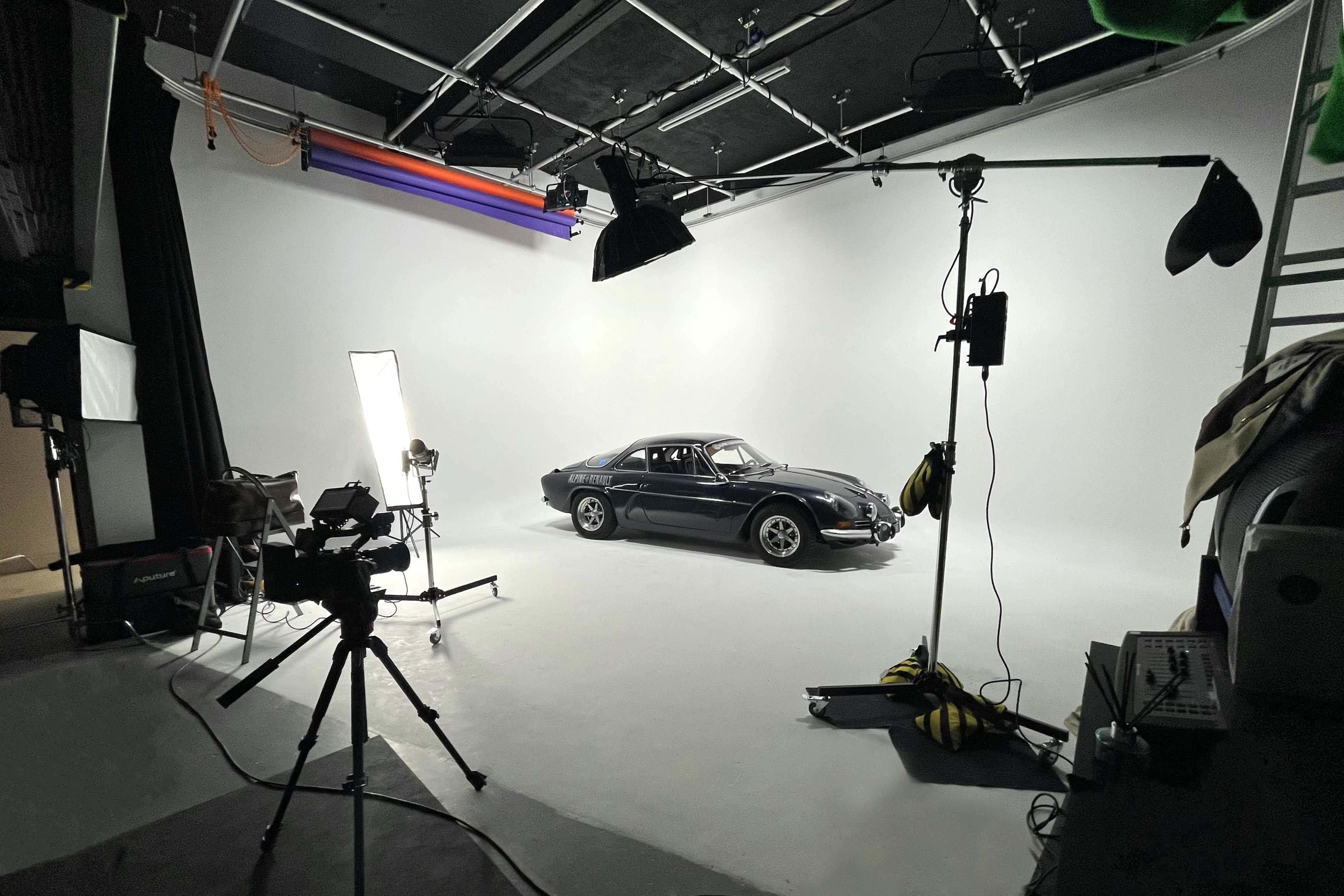 A black car is positioned on a white backdrop in a photography studio, surrounded by lighting equipment and cameras.