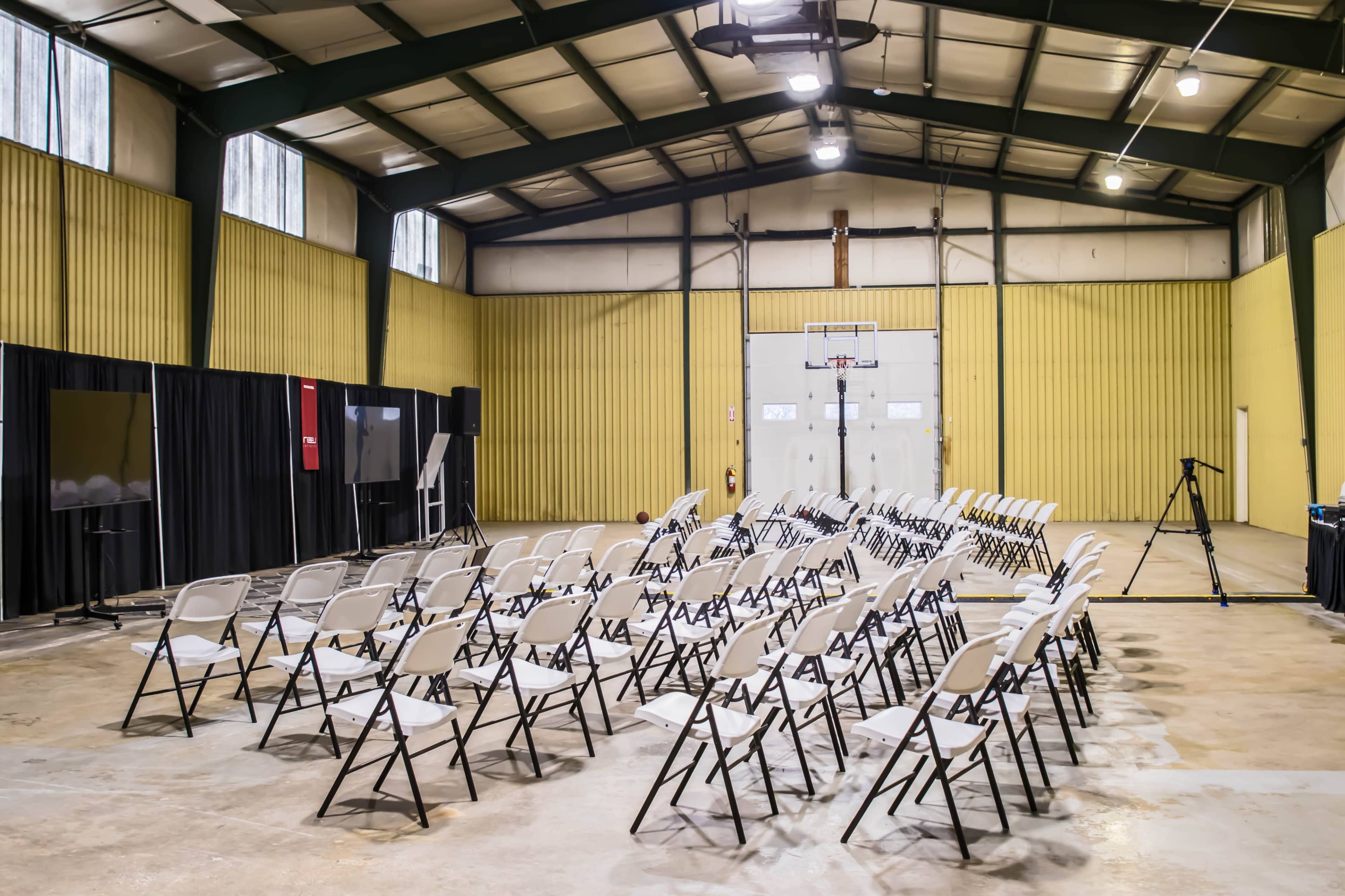 A row of folding chairs is set up in an indoor gymnasium with yellow walls and a basketball hoop in the background.