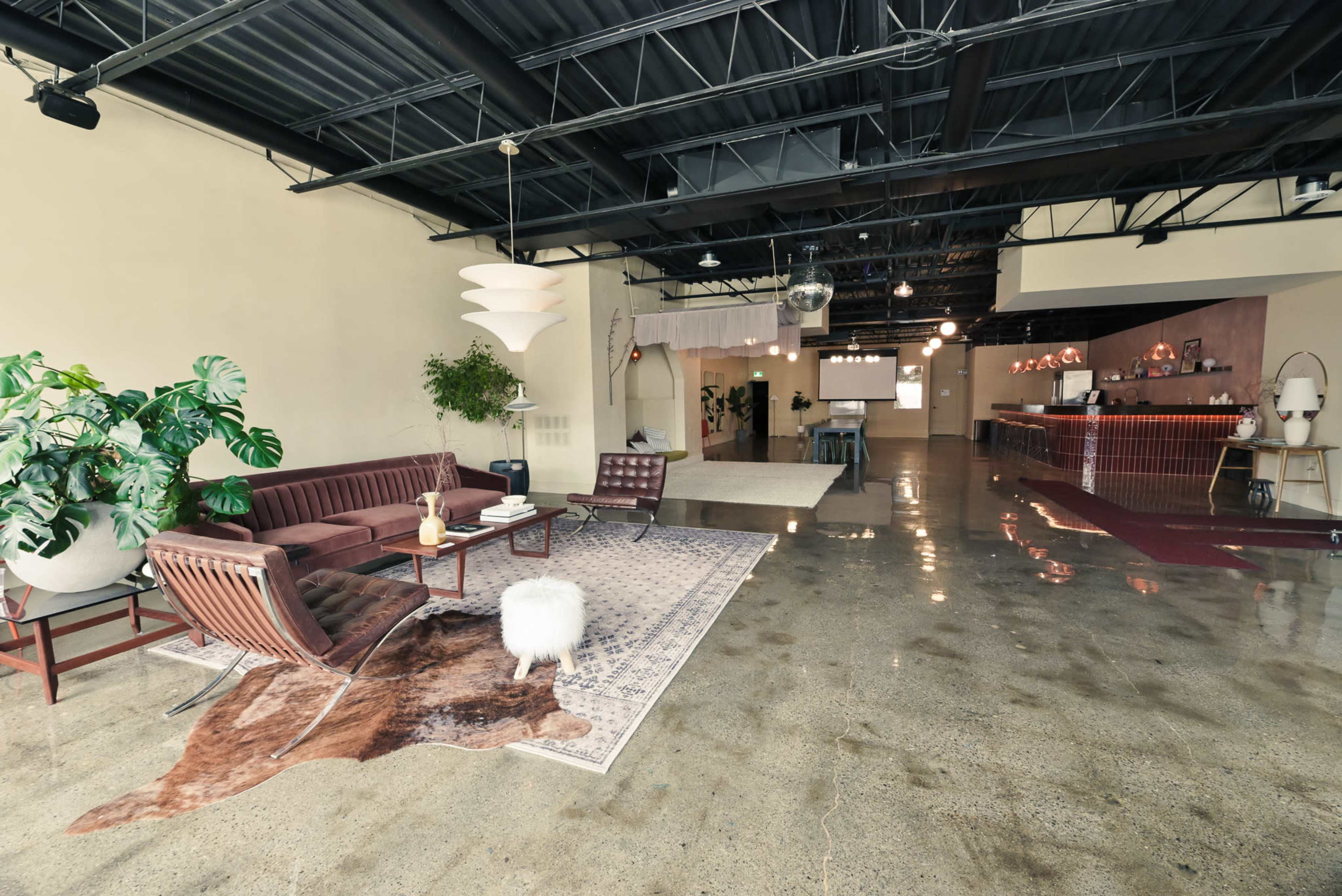 The image shows a spacious, modern indoor area with polished concrete floors, stylish furniture, and ambient lighting, featuring a seating area with a couch and chairs, and a reception or bar area in the background.