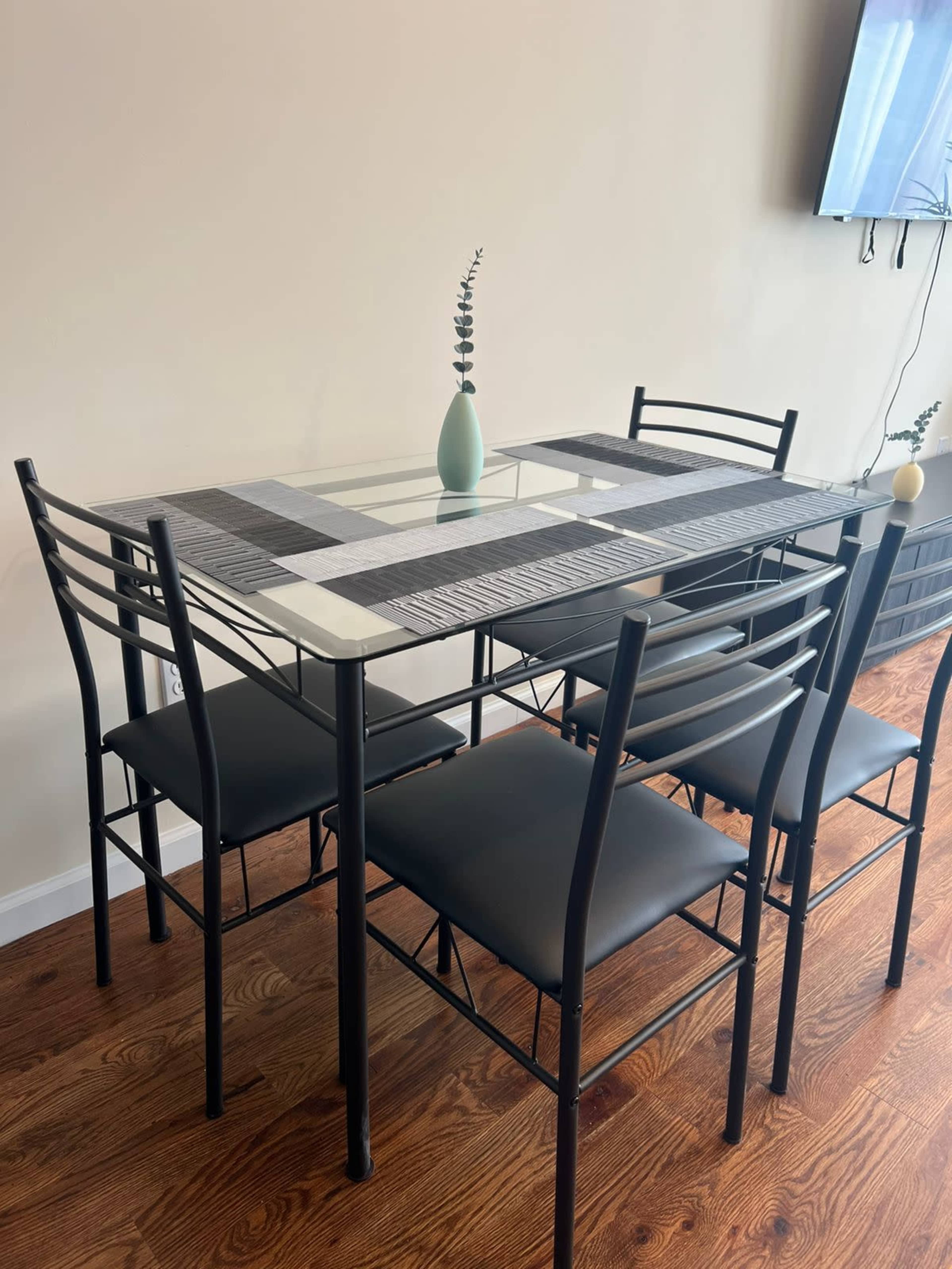 A glass-top dining table with a central runner and five black metal chairs is positioned against a light-colored wall.