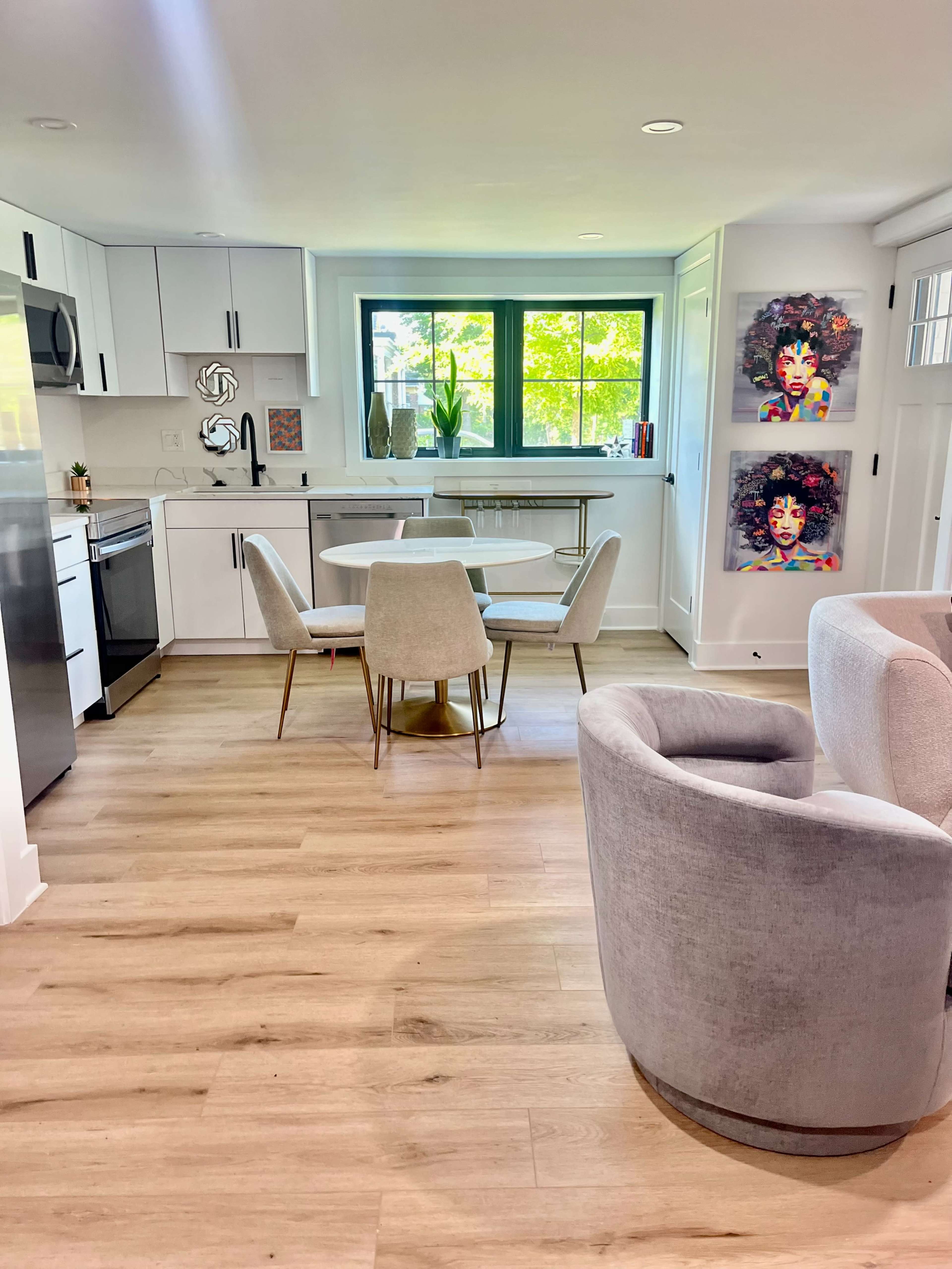A modern kitchen and dining area feature a circular table with four chairs, a gray armchair, and colorful wall art.