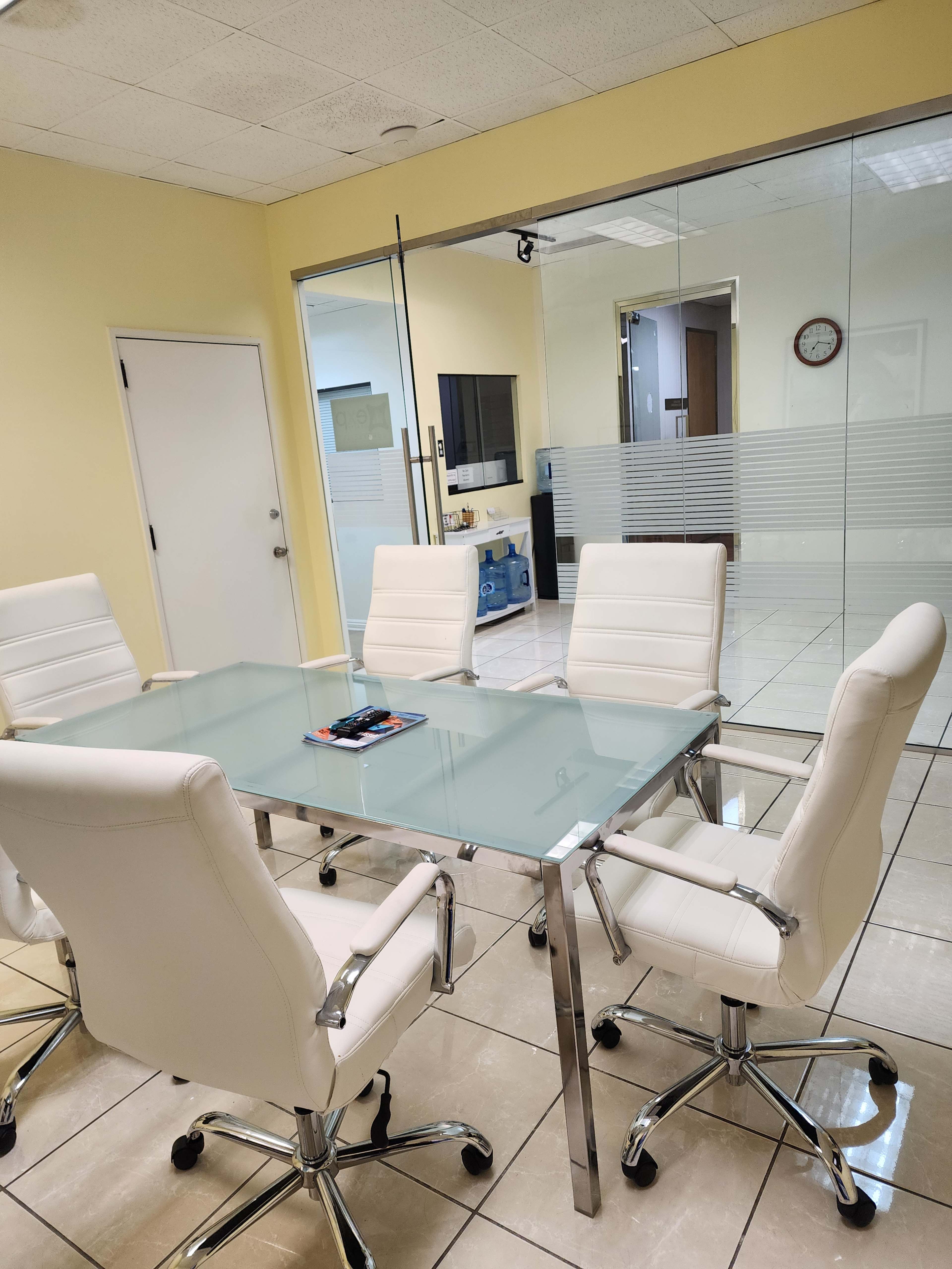 The image shows a conference room with a glass table surrounded by four white leather chairs and a wall of glass, revealing an adjoining space.