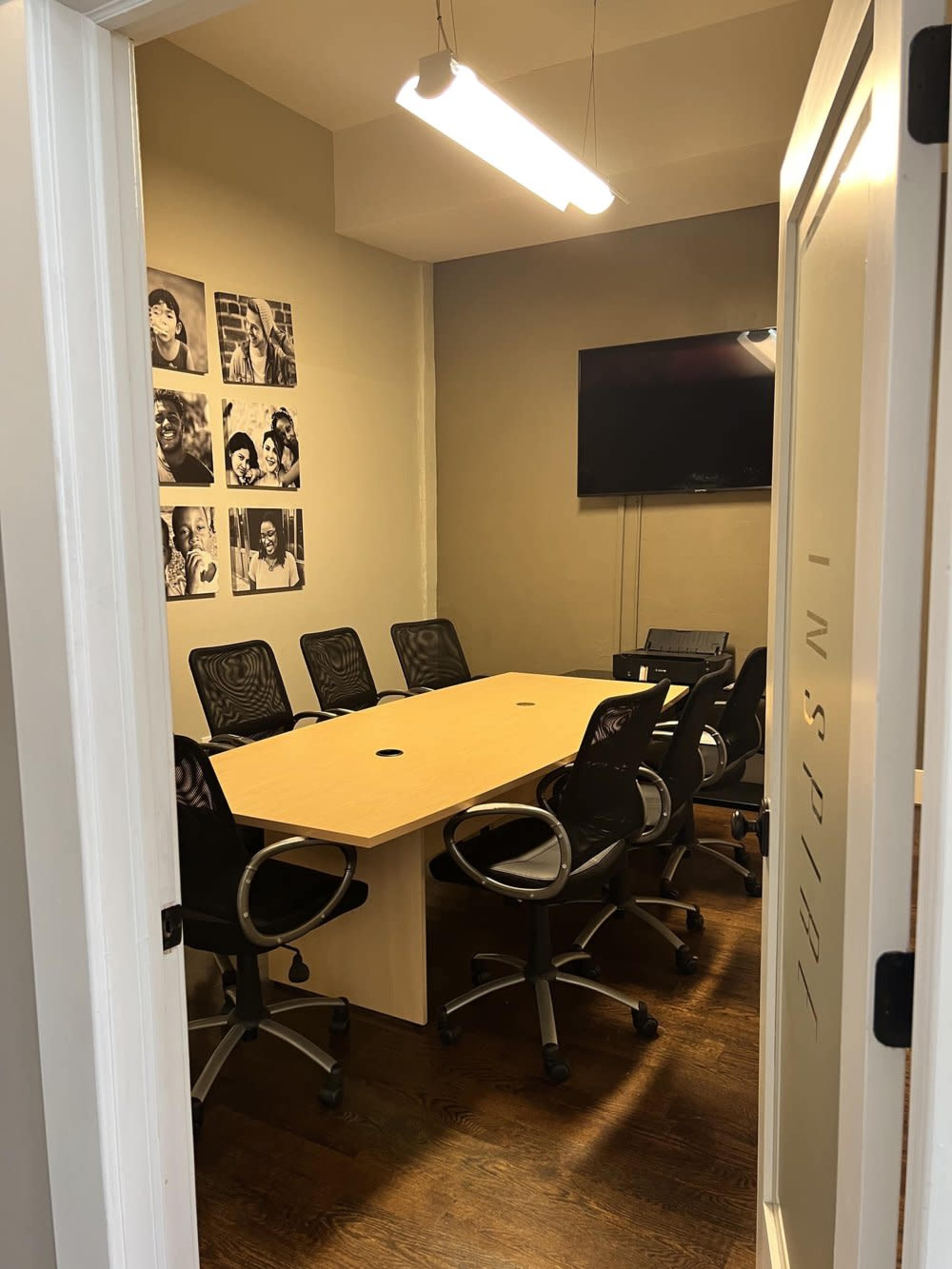 A small conference room with a large wooden table surrounded by six black office chairs, and a wall adorned with framed black-and-white photos.