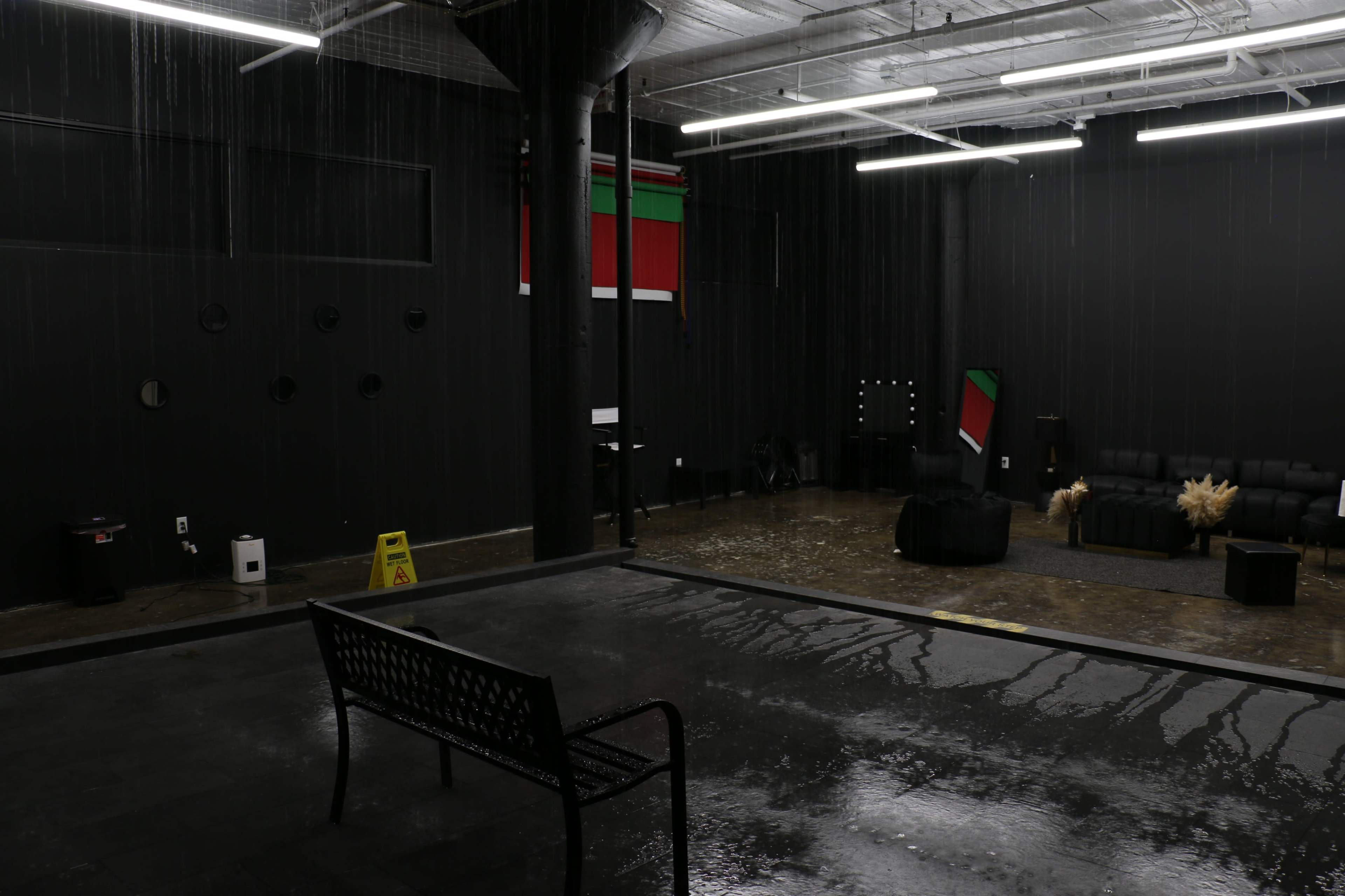 The image shows a spacious room with black walls and flooring, featuring a bench in the foreground and water pooling on the ground from a ceiling leak, while a caution sign is visible nearby.