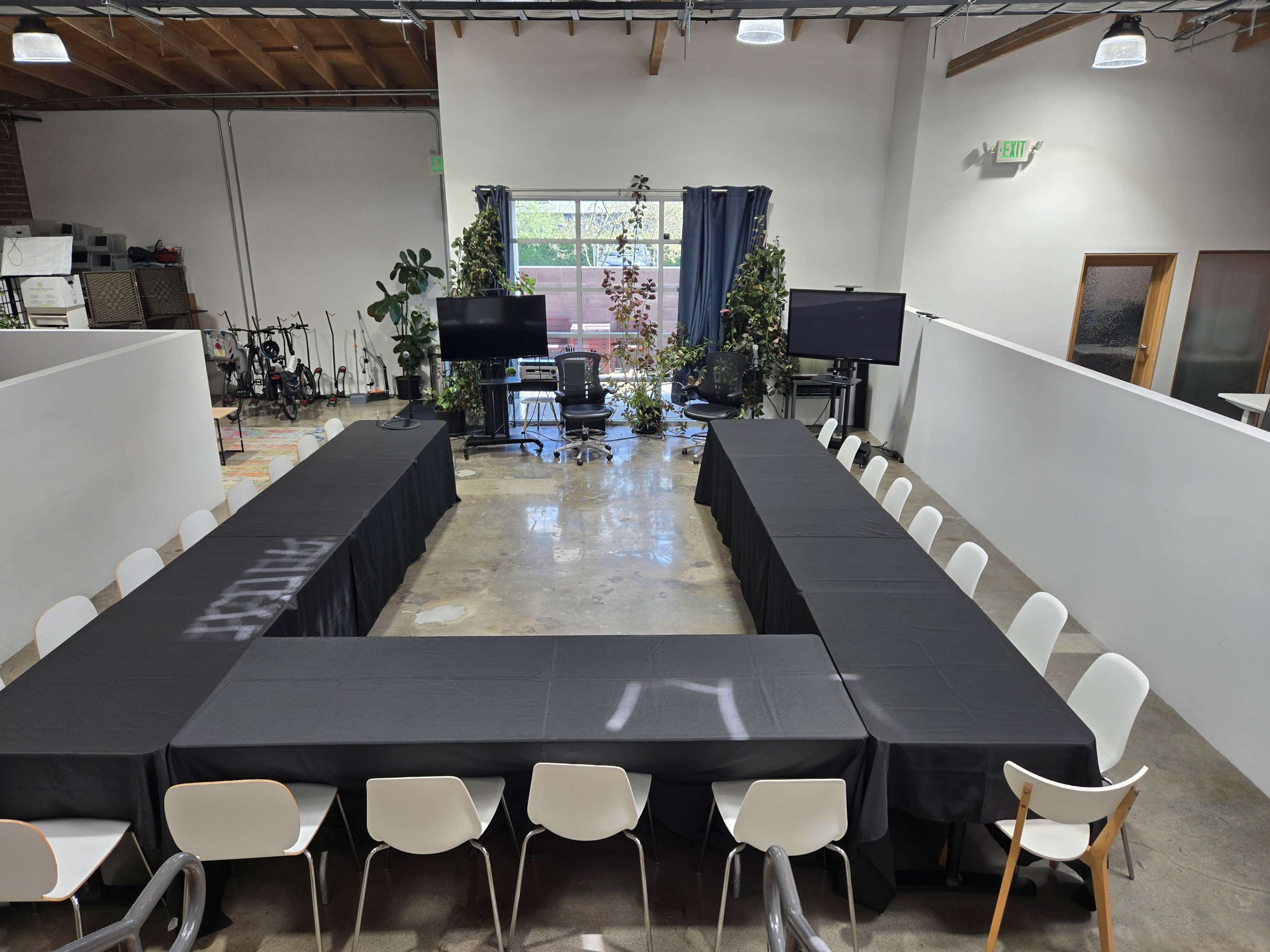The image shows a spacious conference room set up with a U-shaped arrangement of tables covered in black cloth, surrounded by chairs and greenery.