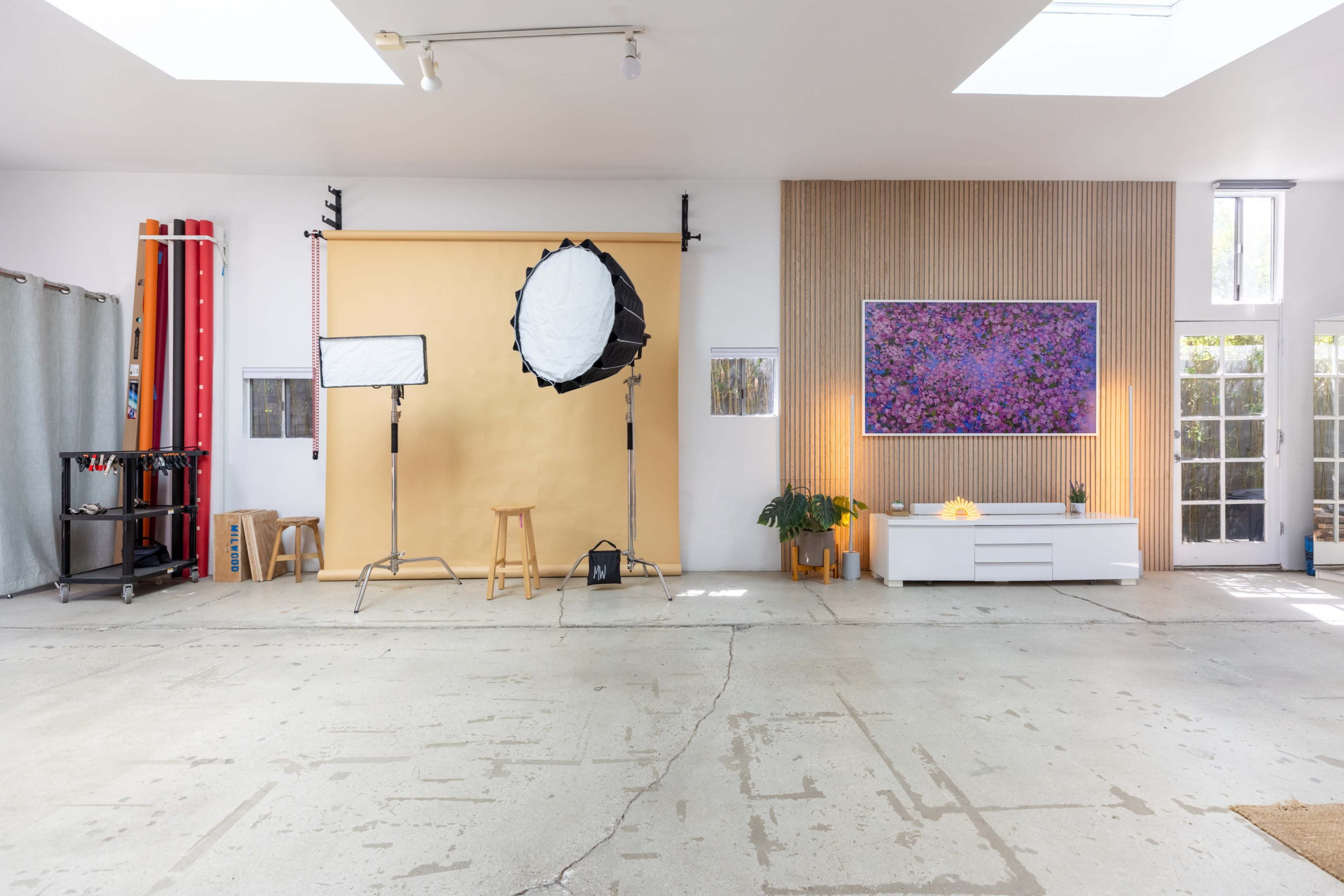 The image shows a photography studio with a yellow backdrop, a light setup, a white cabinet, and a colorful artwork on the wall.
