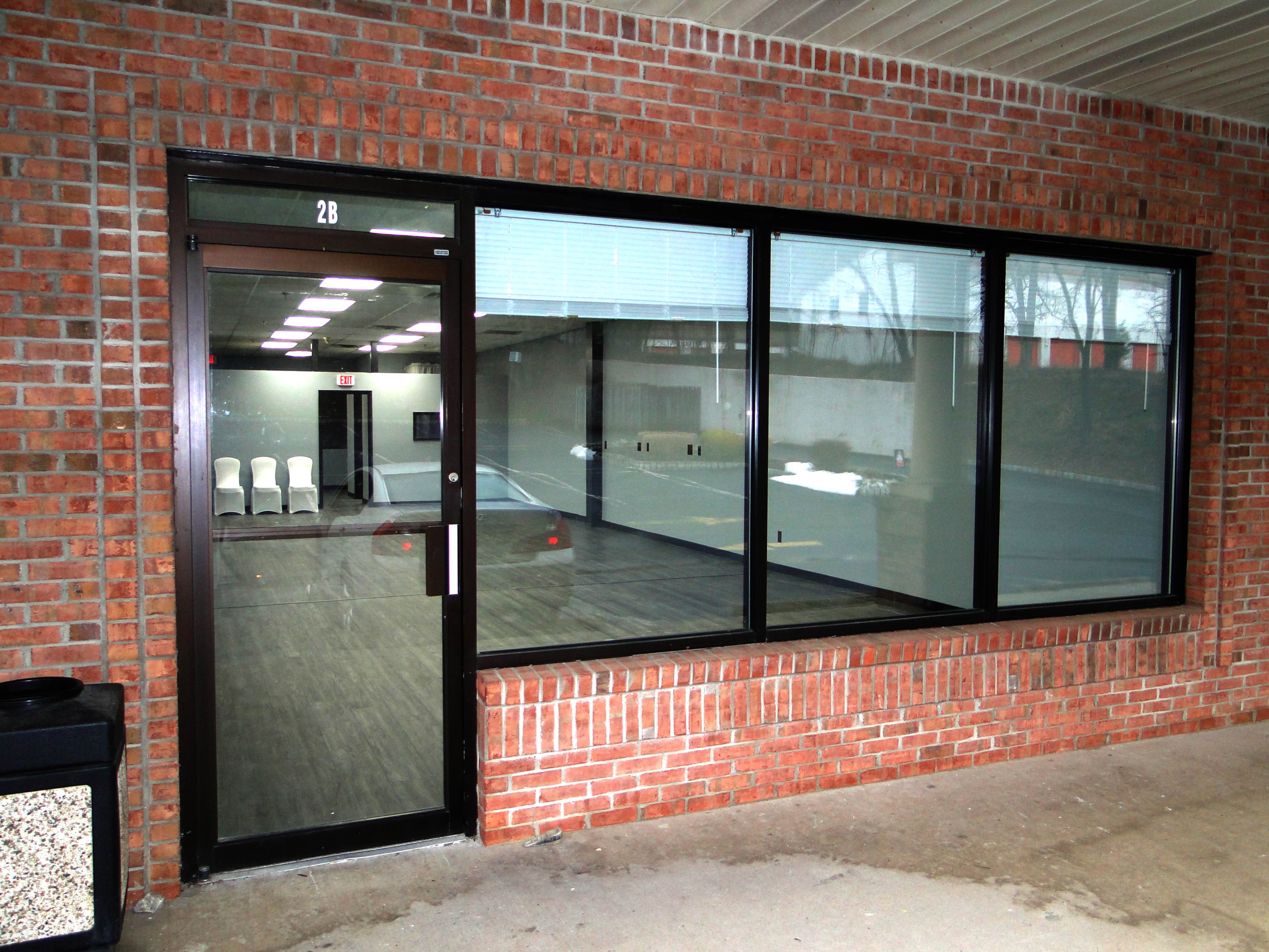 A storefront with large glass windows and a black door, situated against a red brick wall.