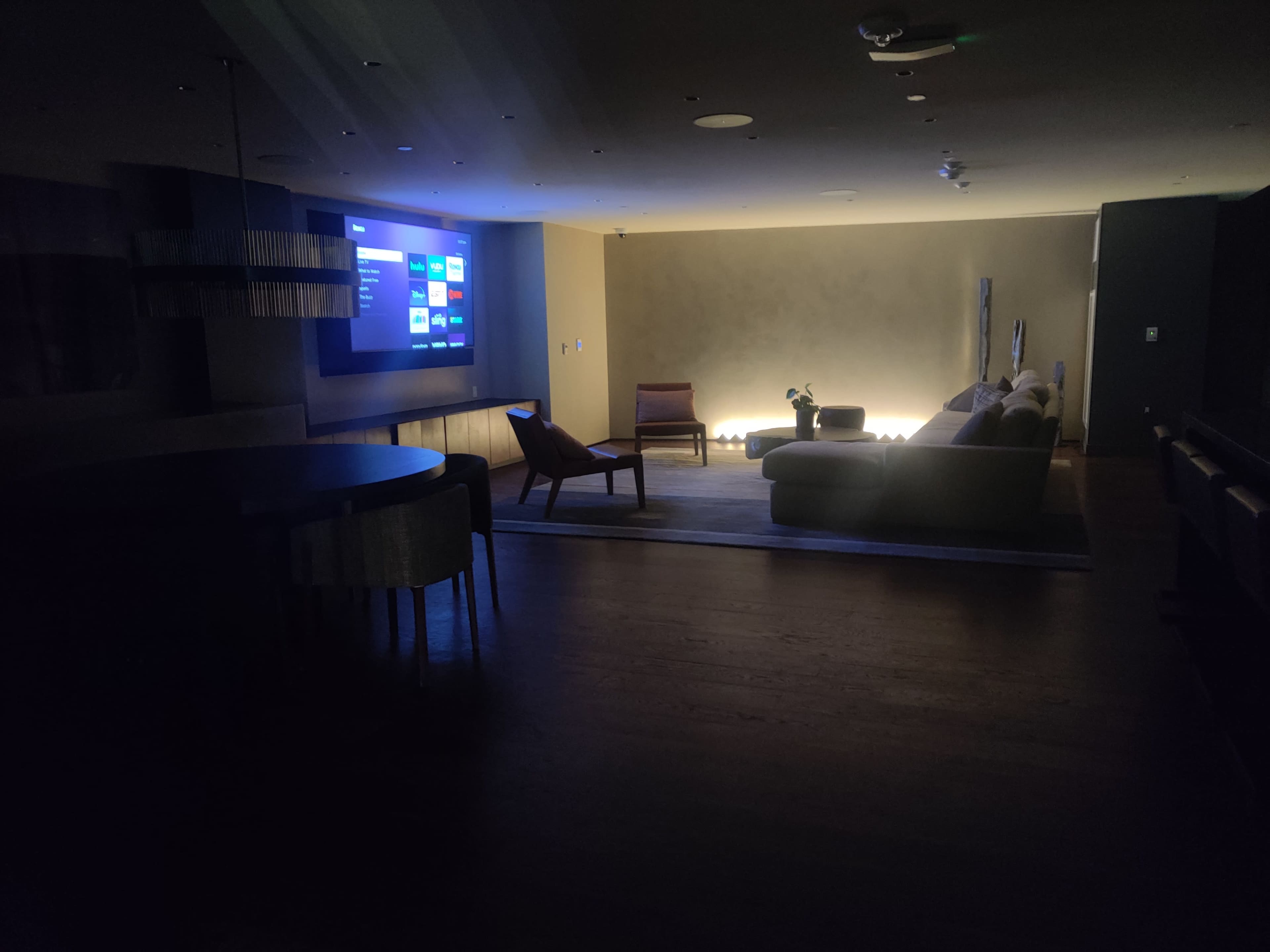 The dimly lit room features a large screen displaying content on one wall, with a sofa and a chair arranged around a low table.