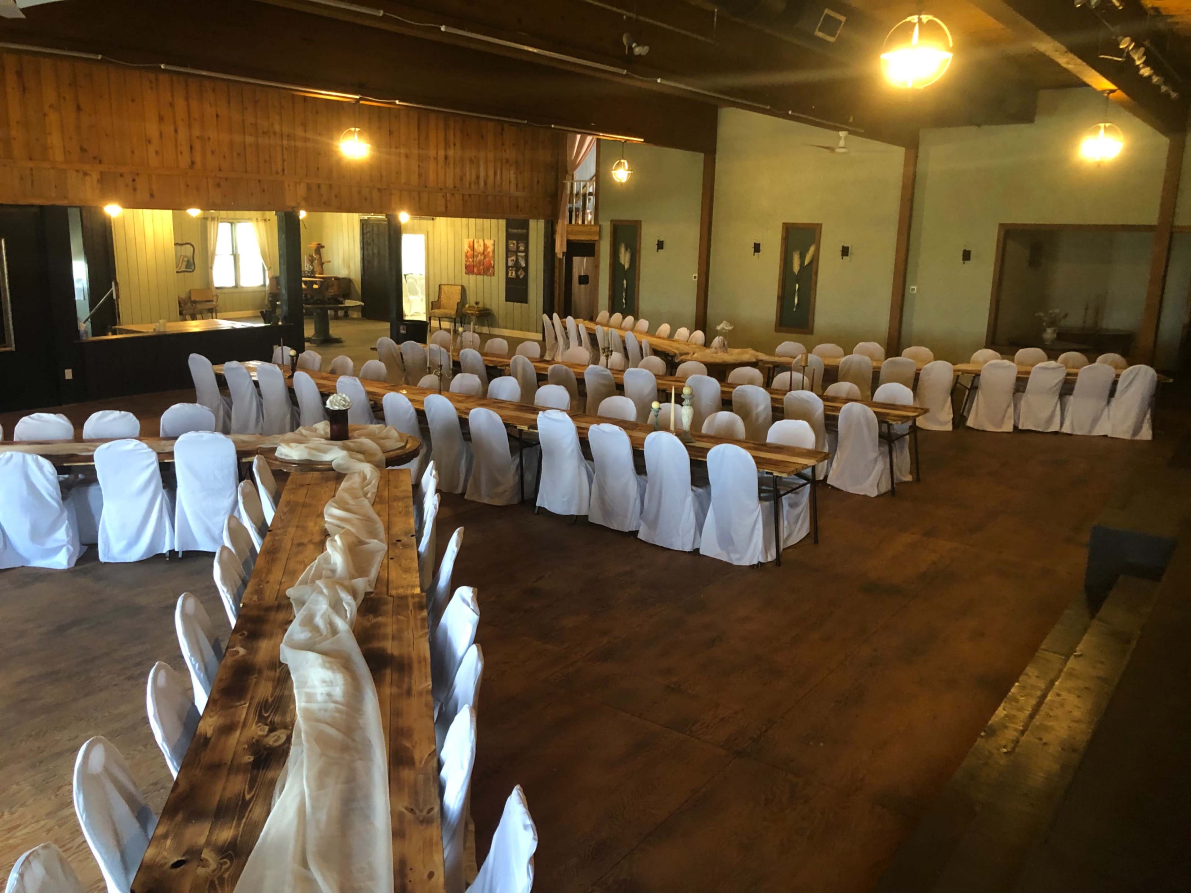 A spacious indoor venue features multiple long tables arranged with white chair covers and a central decorative table runner.
