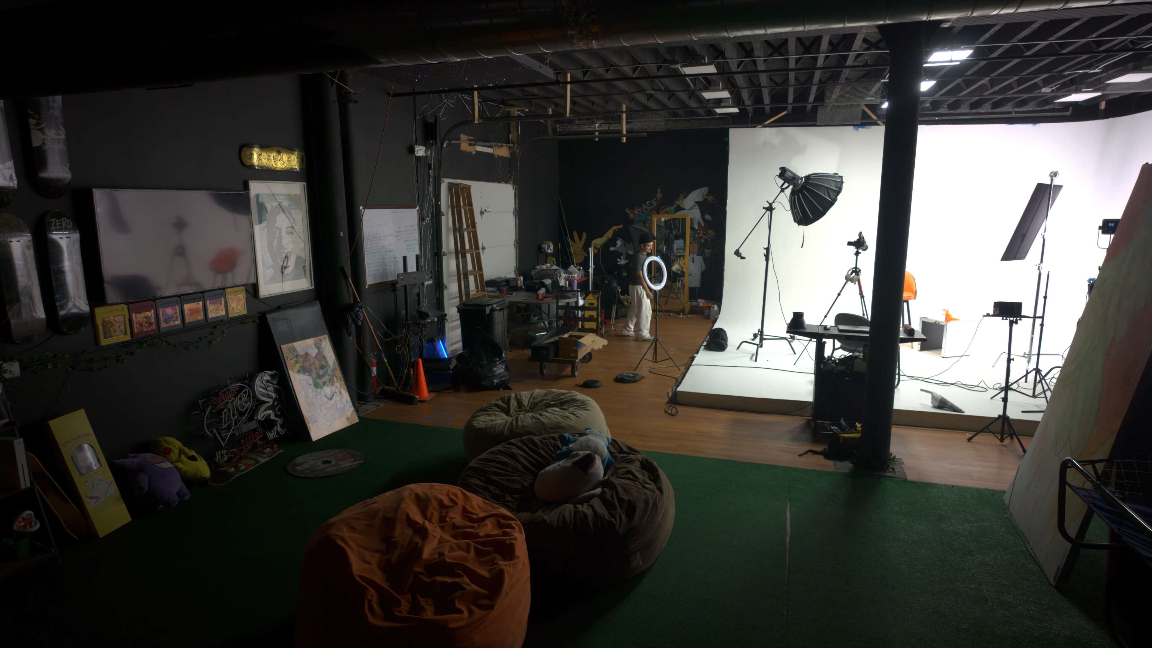 The image shows an indoor studio space featuring photography equipment, lighting setups, a blank backdrop, and various props, with bean bags and a carpeted floor.