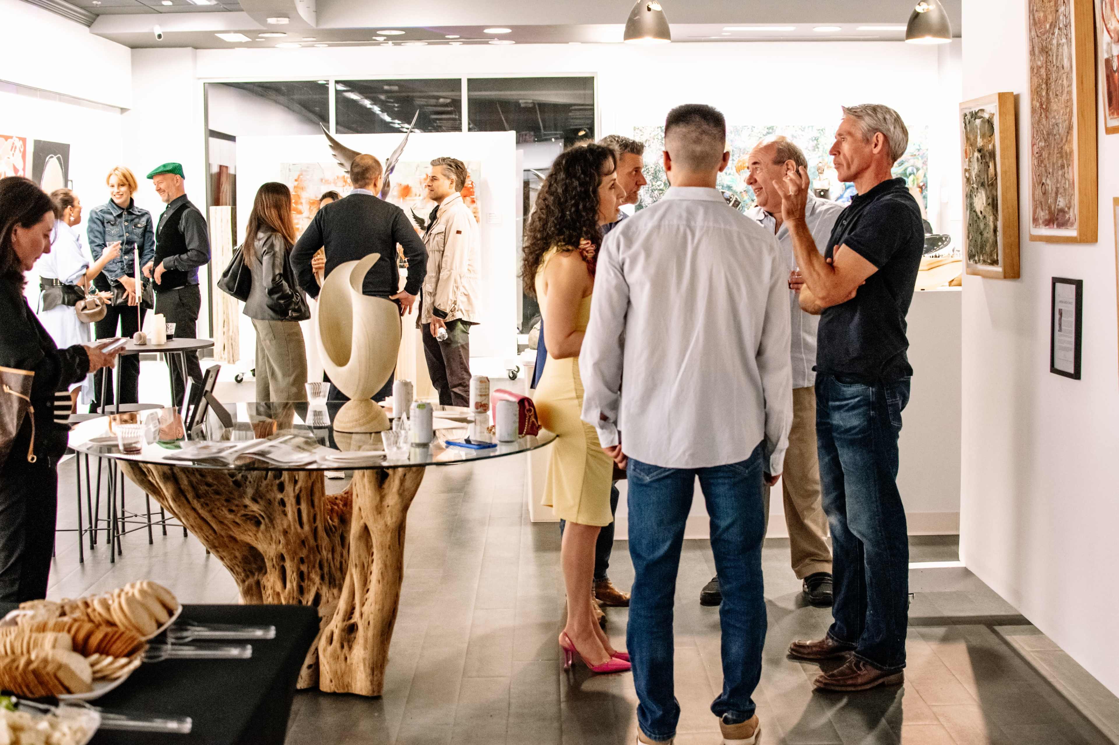 A group of people converse and mingle at an art gallery opening, surrounded by various artworks displayed on the walls.
