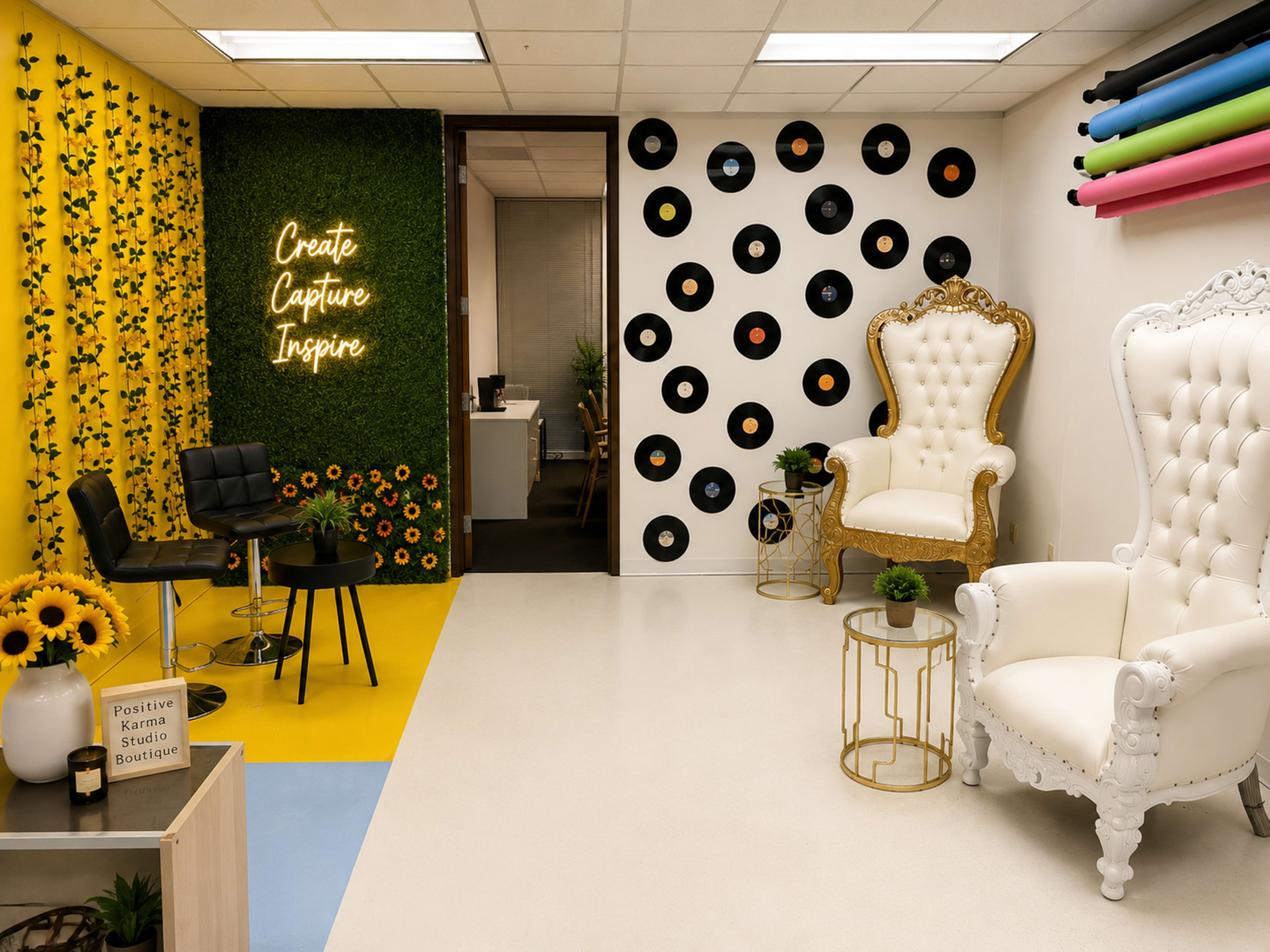 A brightly colored room features floral and vinyl record wall decor, elegant white chairs, and a neon sign that reads "Create Capture Inspire."