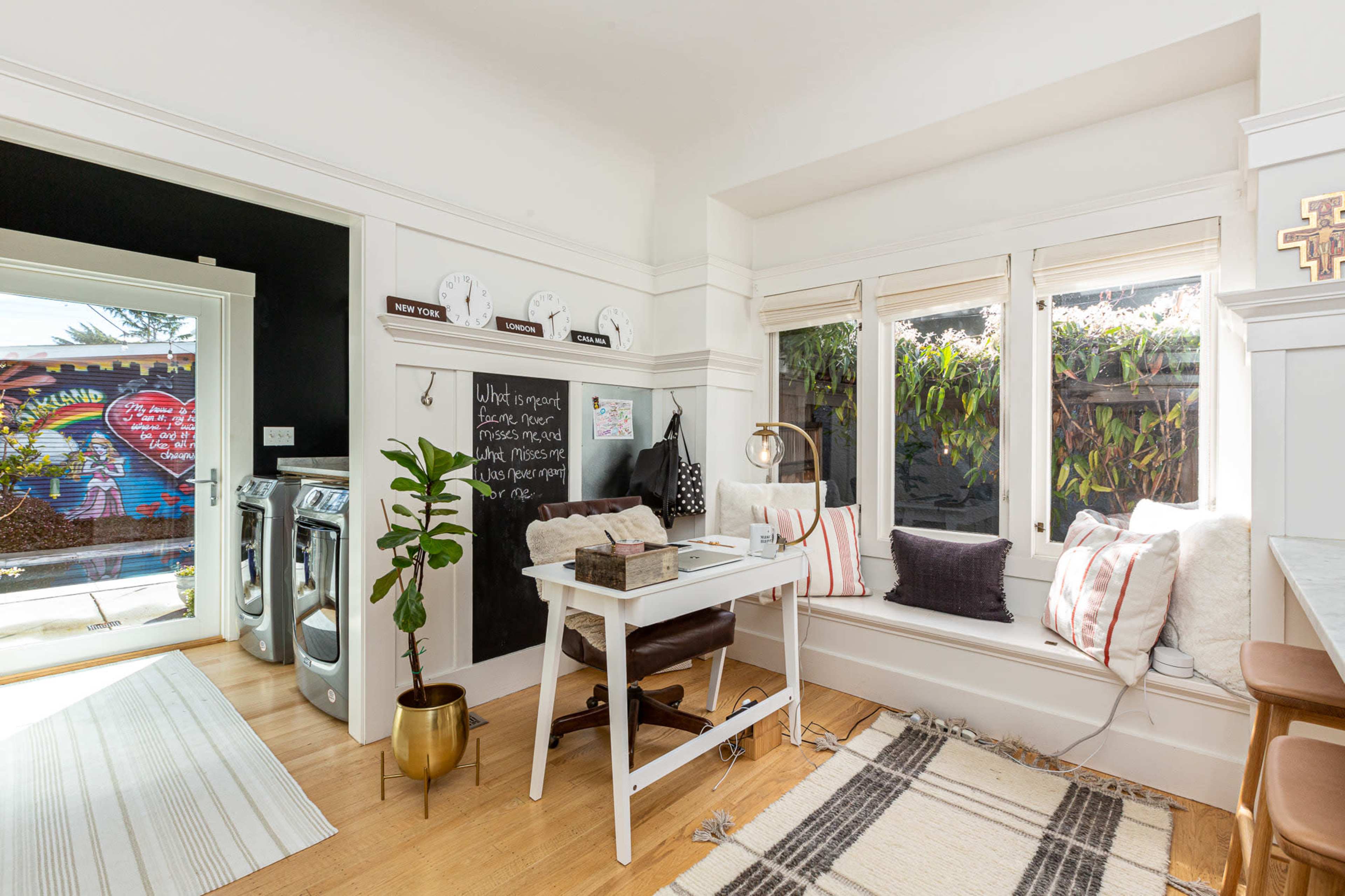 A bright, minimalist home office features a white desk beside large windows, a potted plant, and a wall with chalkboard paint.