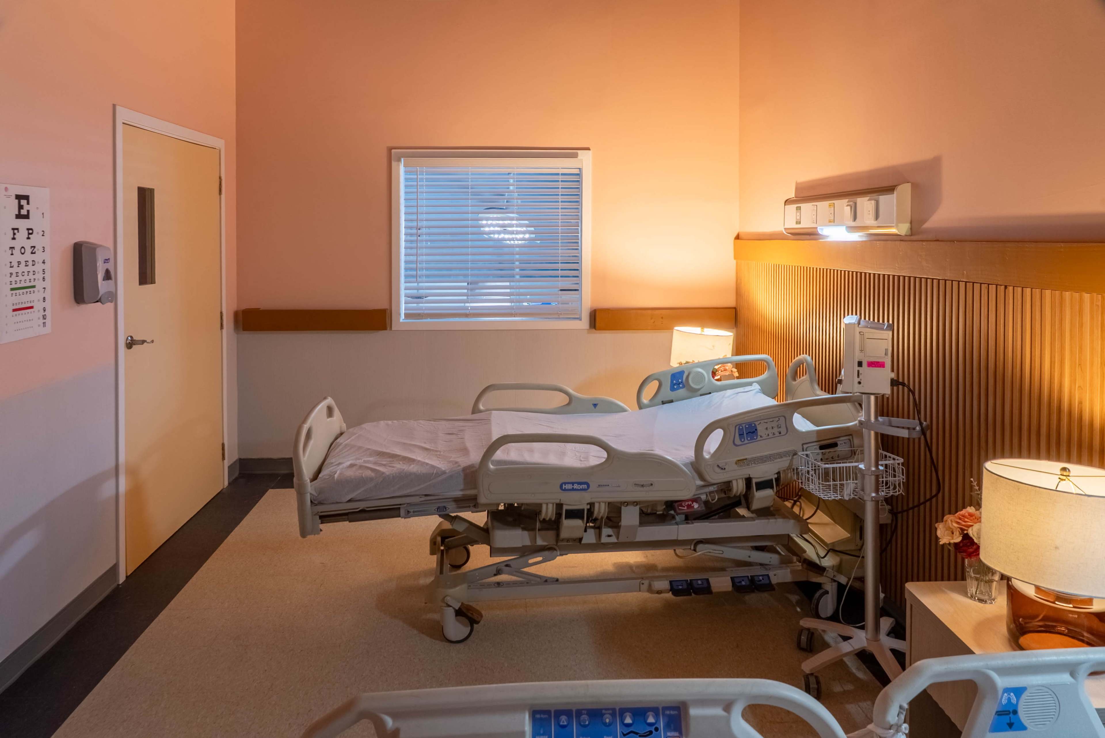 The image shows a hospital room with a hospital bed, a bedside lamp, a nightstand, and a window.